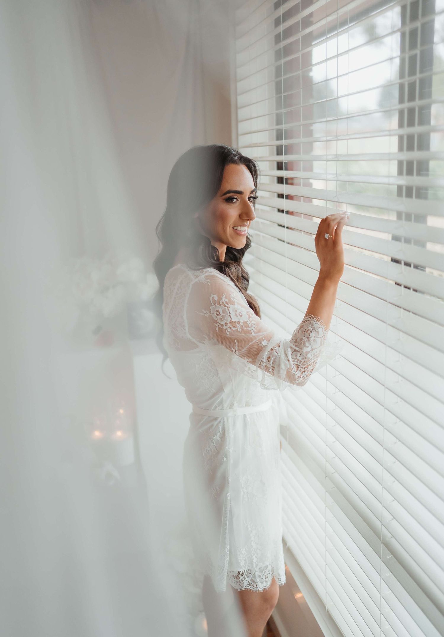 Figure in white lace robe stands by window with soft morning light creating ethereal atmosphere.