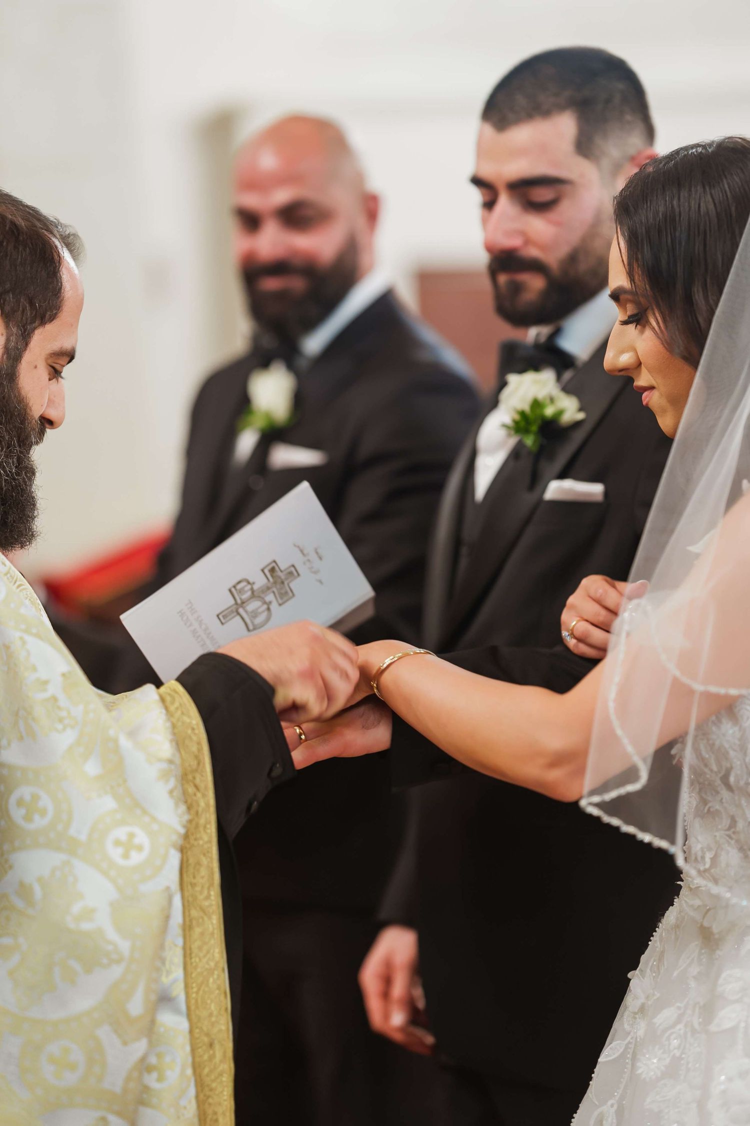 A wedding ceremony showing hands being joined during the exchange of vows, with attendants in dark suits looking on.
