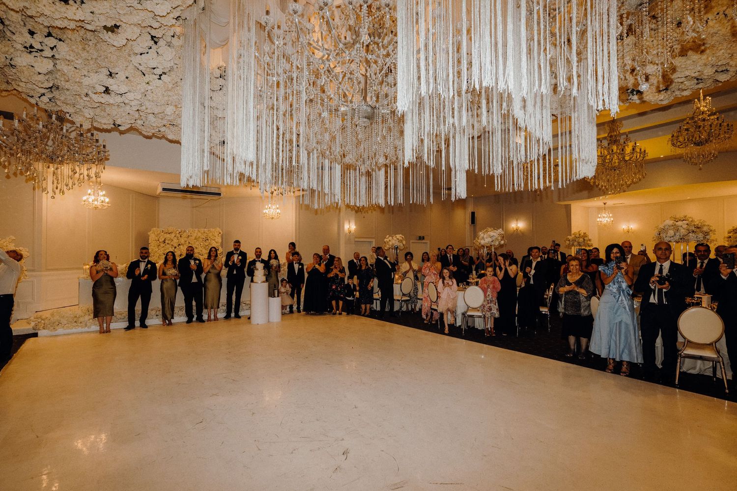 Wedding guests stand in an ornate gilded ballroom with crystal chandeliers during an elegant reception.