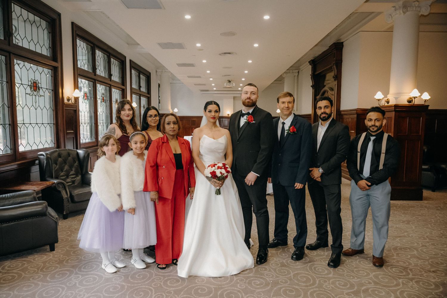Wedding party poses indoors, bride in white gown with bouquet, groom in black suit, guests in formal attire.