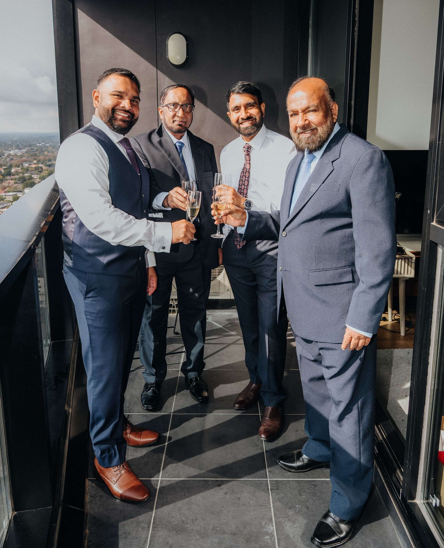 Four businessmen in suits and formal attire toast with glasses on a high-rise balcony overlooking a city view.