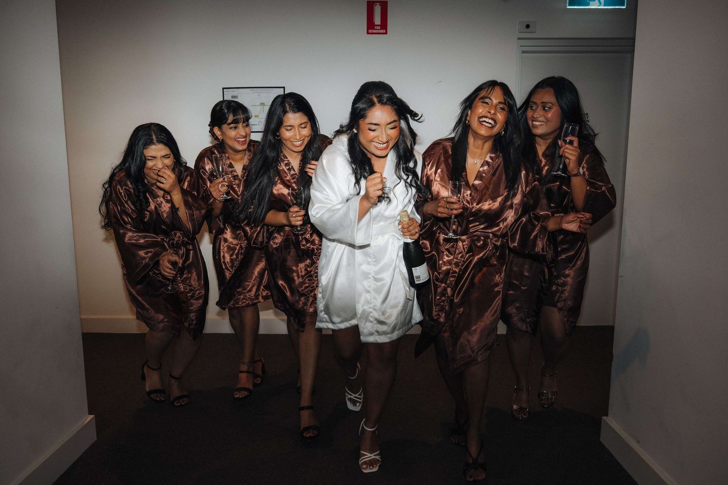 Group of bridesmaids in matching brown satin robes pose with the bride in white robe in a hallway setting.