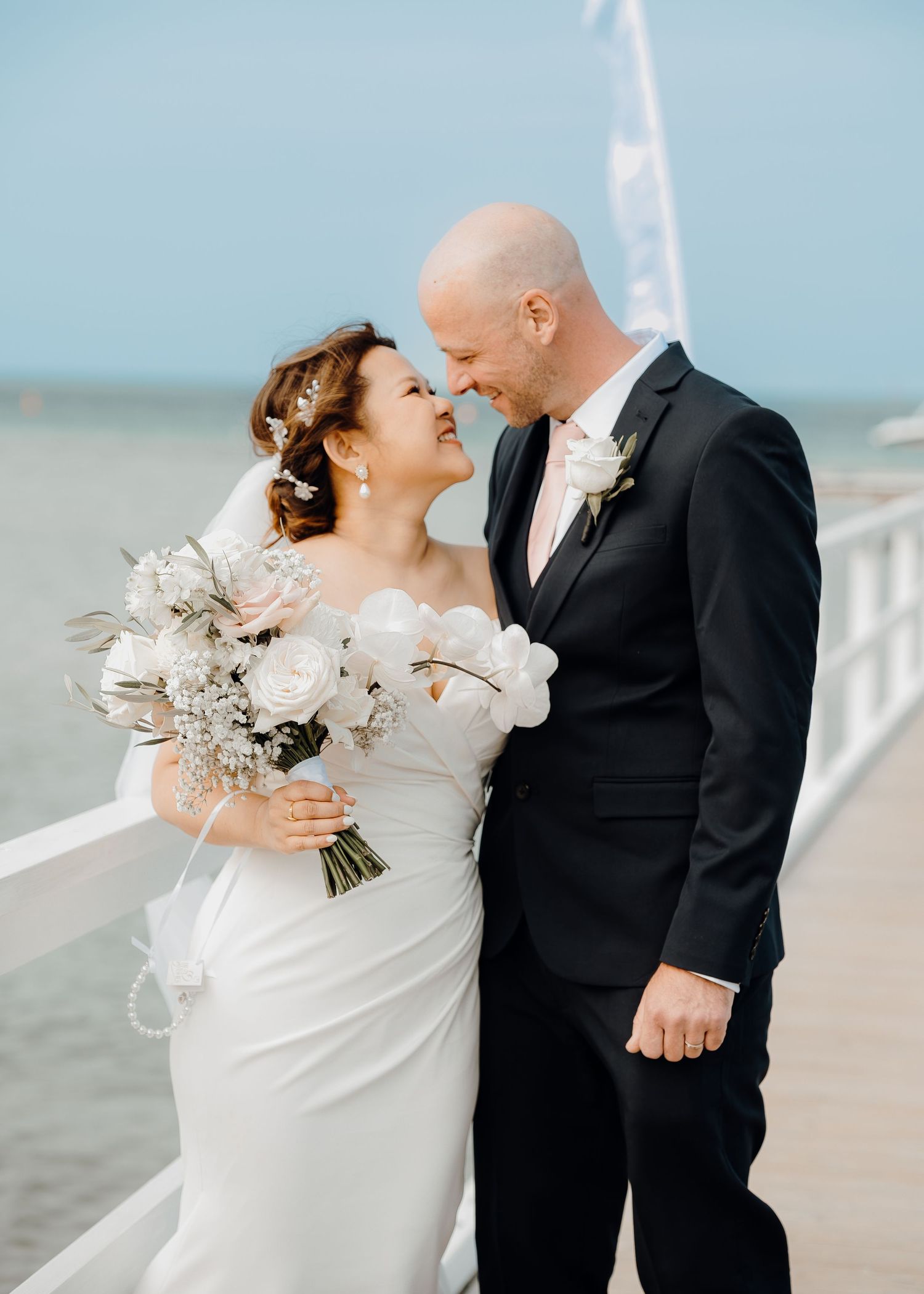 Bride and groom share a romantic moment on a seaside pier, bride holding white bouquet in elegant wedding attire.