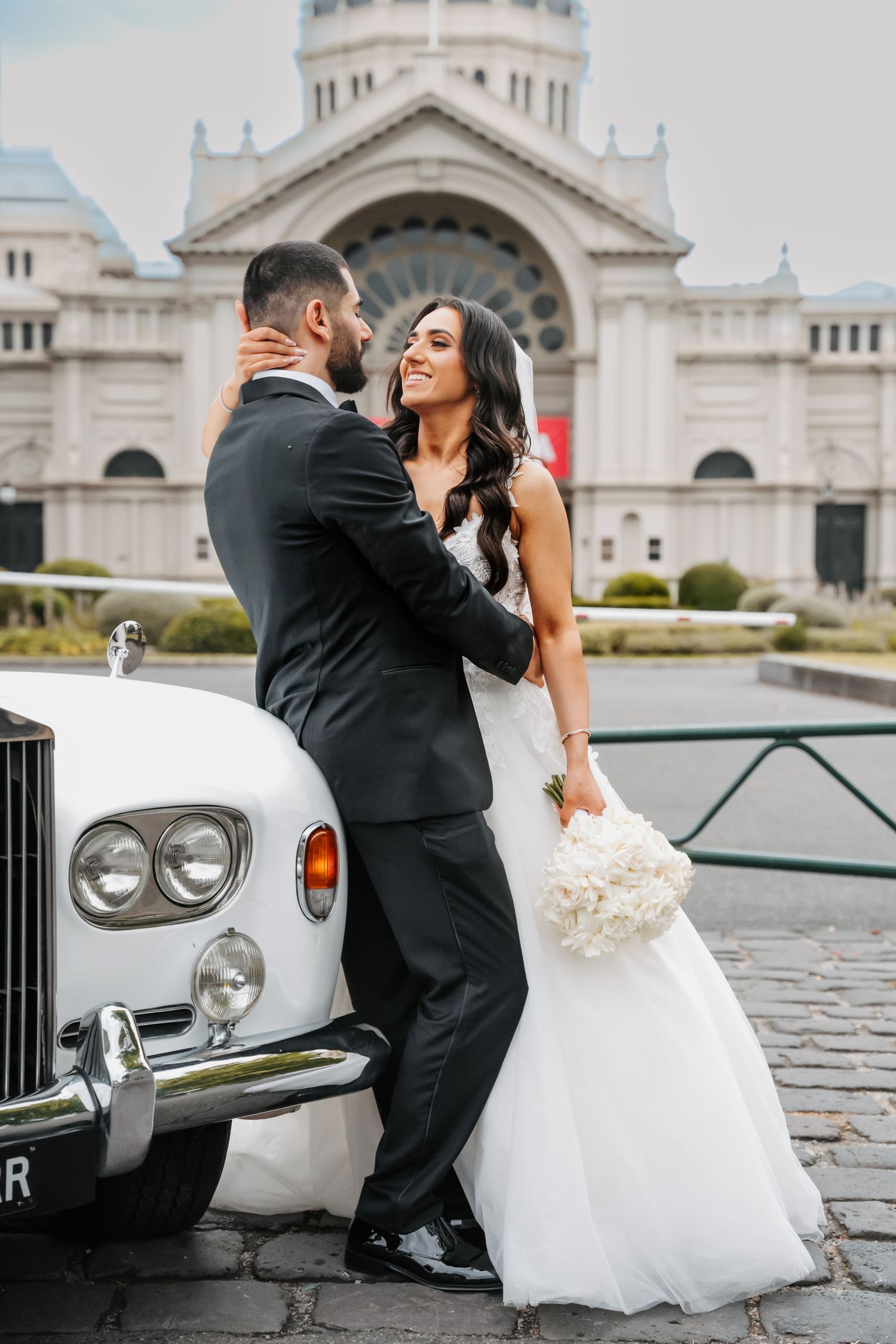 A romantic wedding photo of a couple posing next to a vintage Rolls Royce in front of an ornate cathedral building.