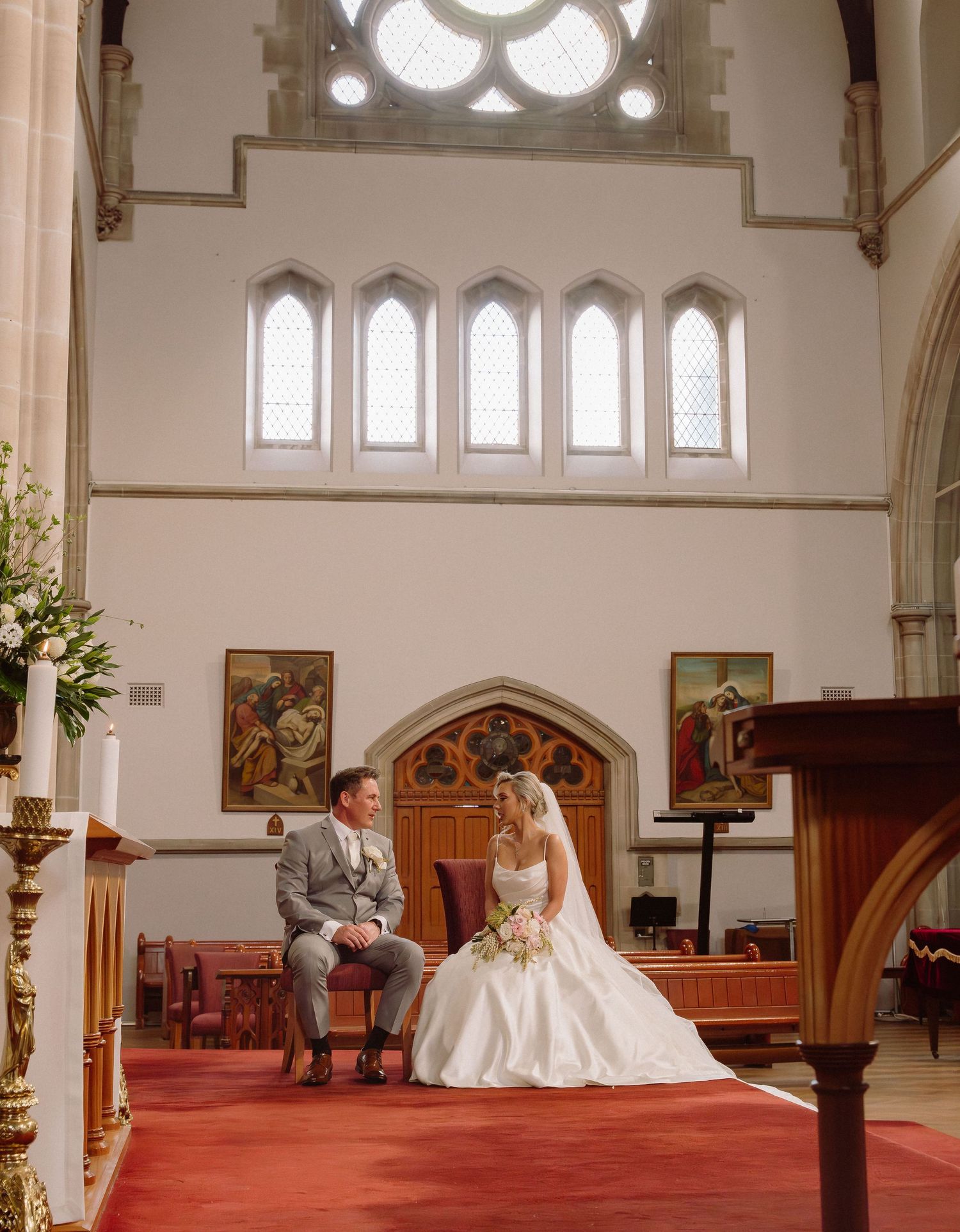Interior of a church with Gothic windows and red carpet, decorated with floral arrangements for a wedding ceremony.