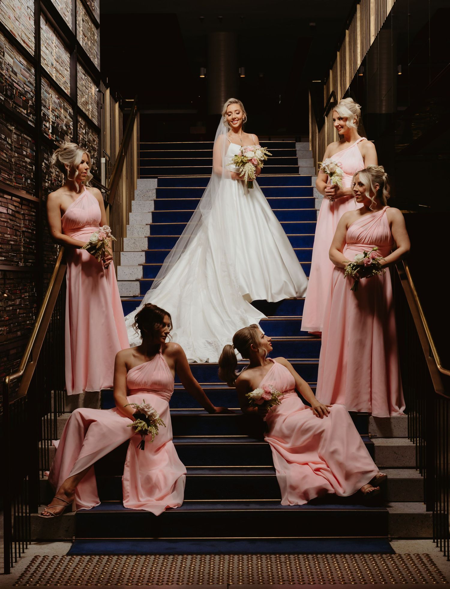 A wedding party poses on elegant dark stairs with bridesmaids in pink dresses and a bride in a white gown.