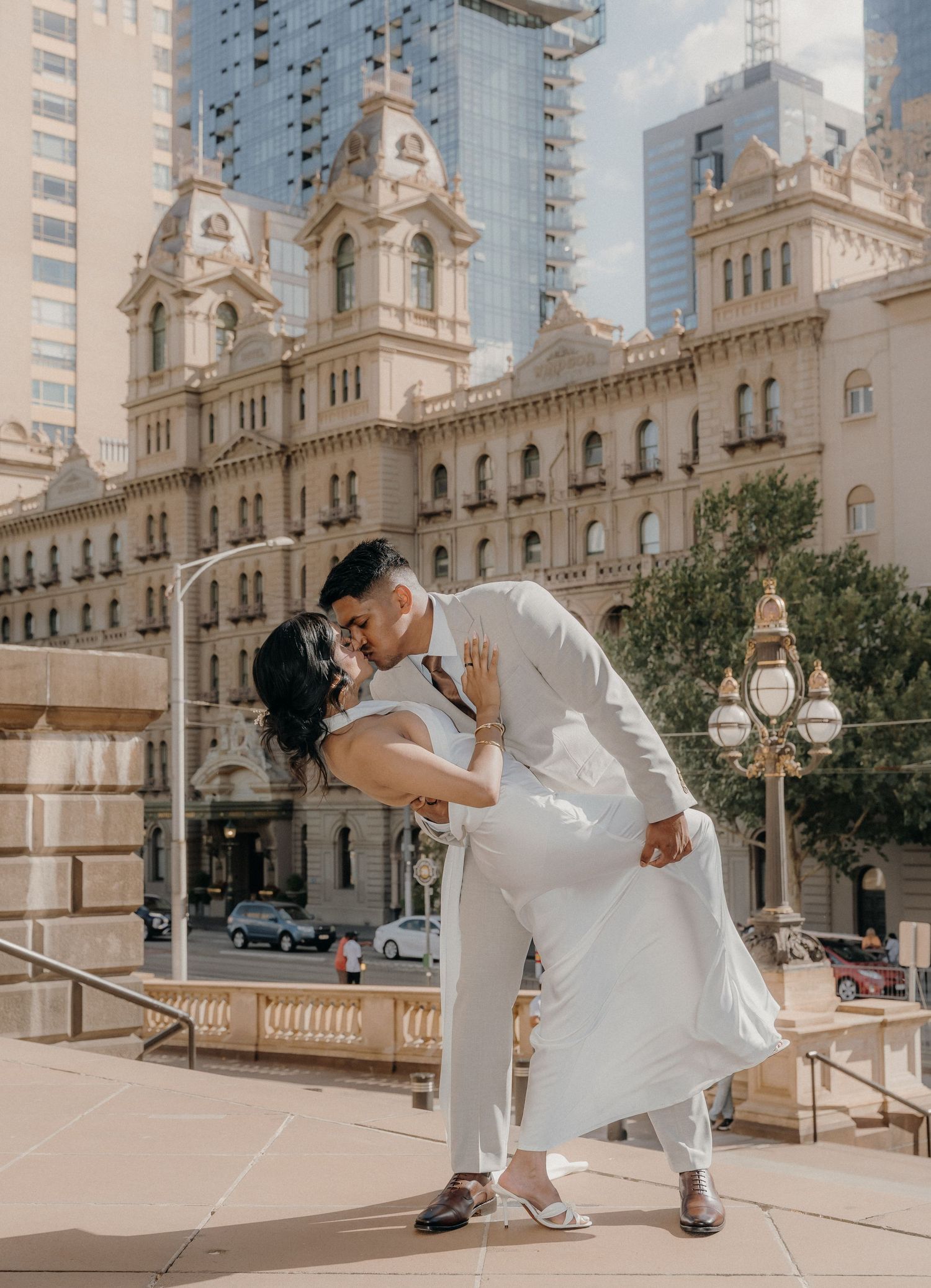 A romantic wedding couple dips and kisses in front of an elegant historic building with ornate architecture.