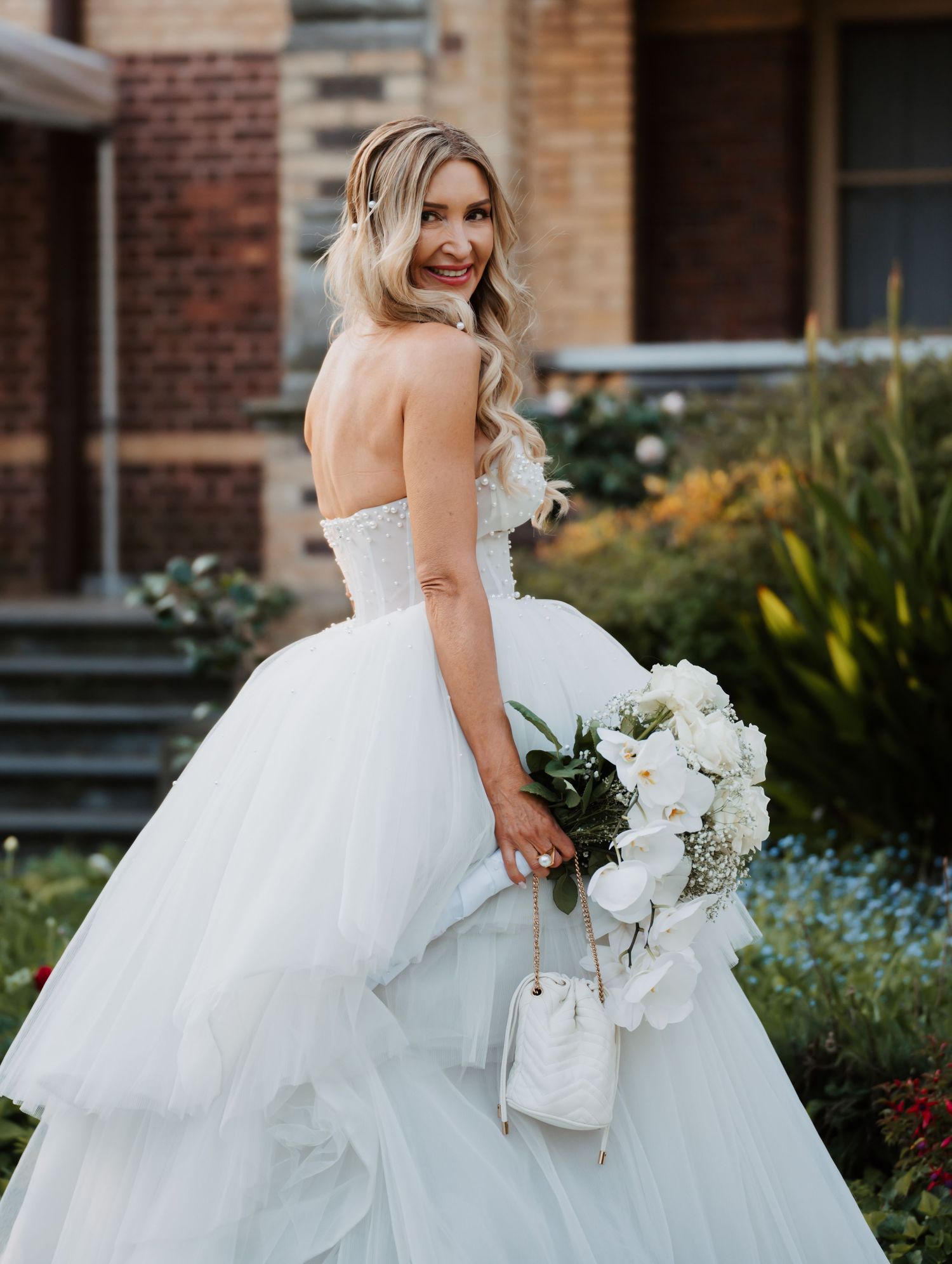 A bride in a strapless white ballgown with a full tulle skirt holds a bouquet of white flowers against a brick backdrop.