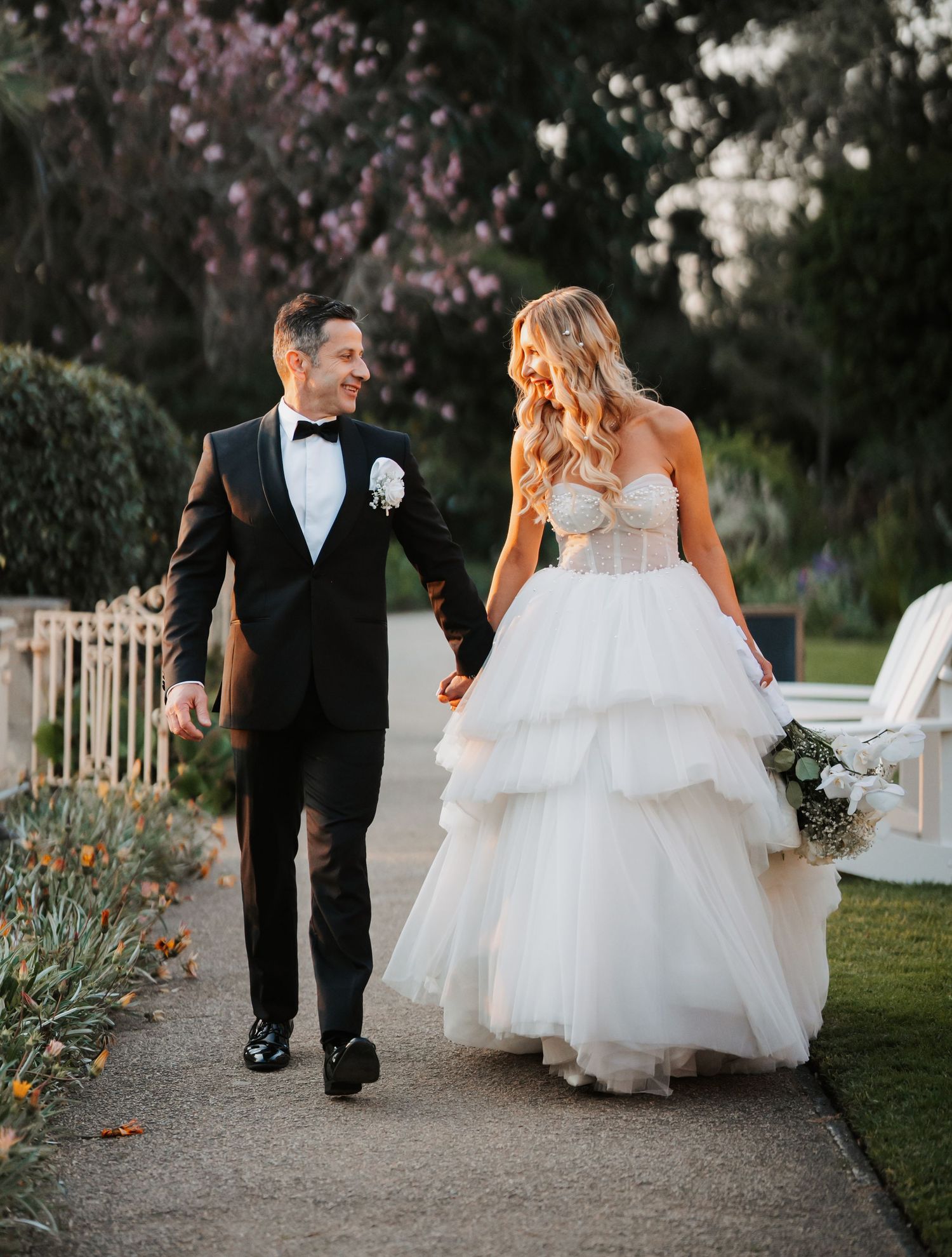 A couple in wedding attire walks together on a garden path with blooming cherry blossoms in the background.