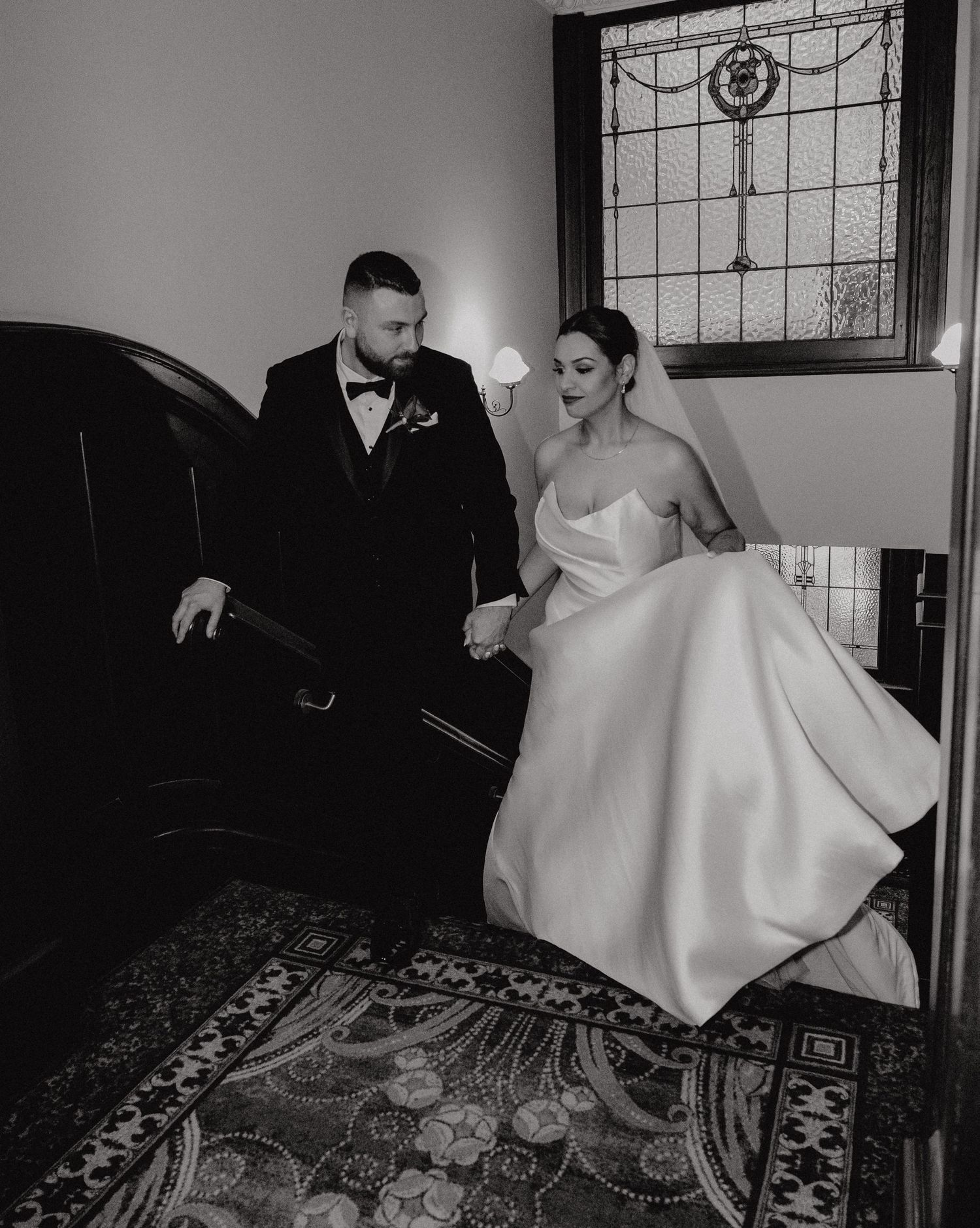 Black and white wedding photo of bride in strapless gown and groom in tuxedo holding hands on ornate staircase.