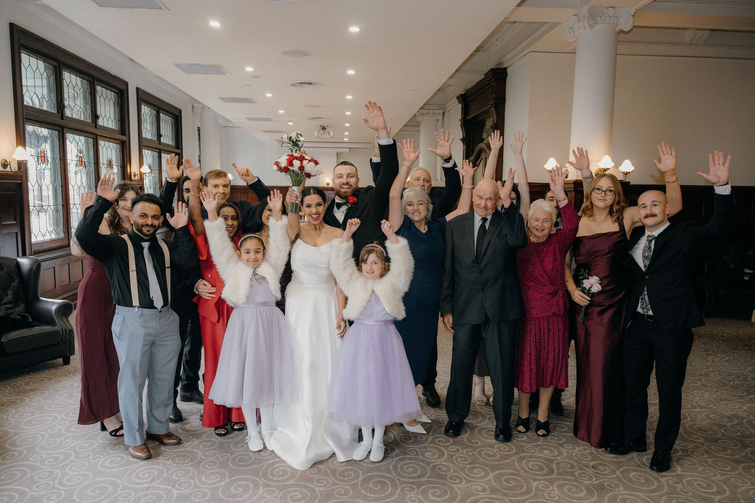 Wedding group photo with bride, groom, and guests cheering with raised arms in elegant venue.