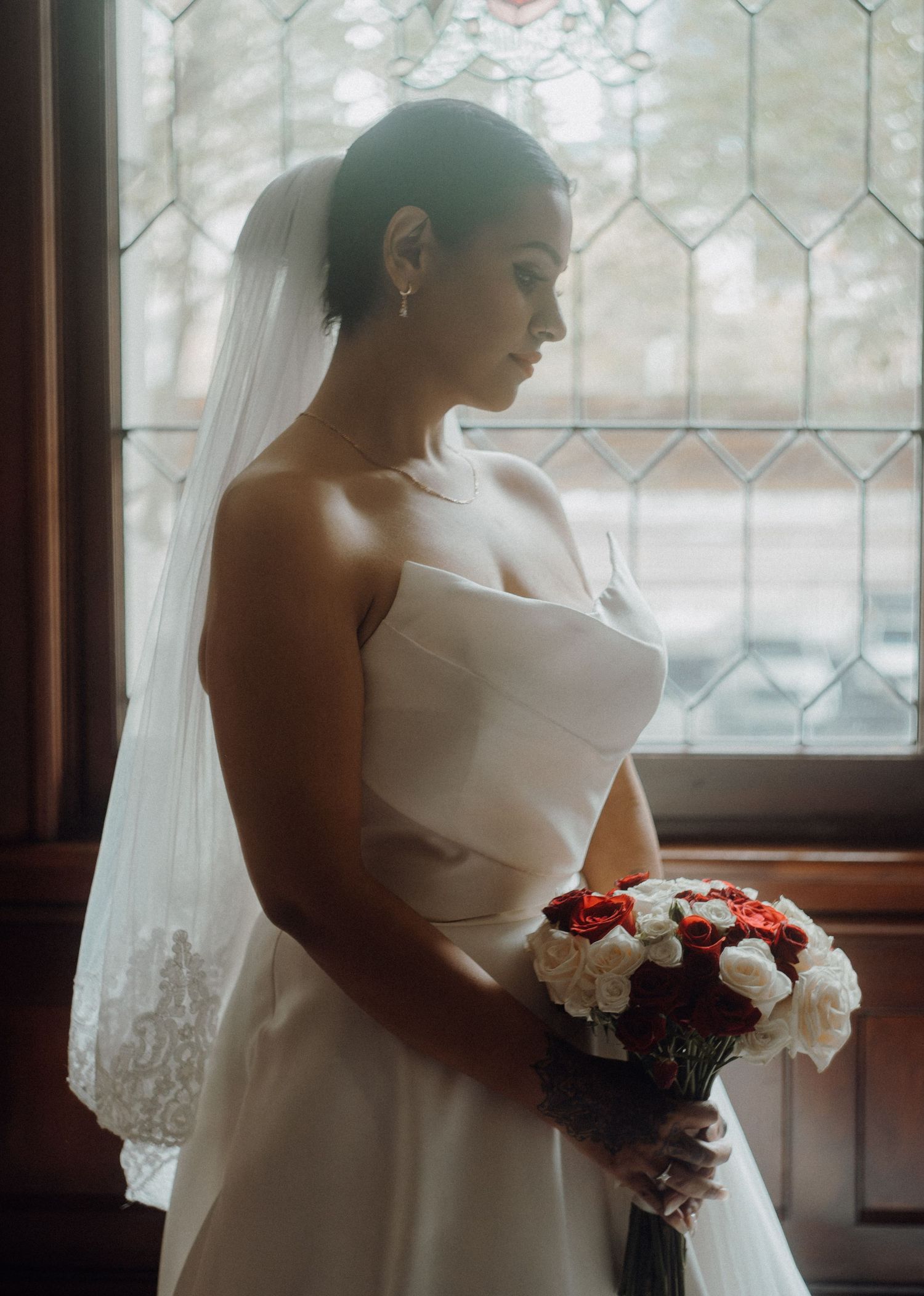 Bride in strapless white gown and veil holds red and white rose bouquet by stained glass window.