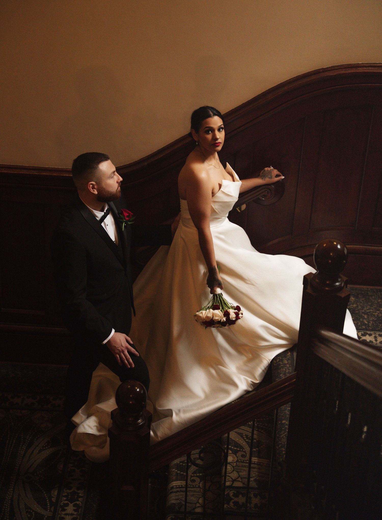 Bride in elegant satin gown holds bouquet on grand staircase beside groom in black tuxedo at wedding venue.