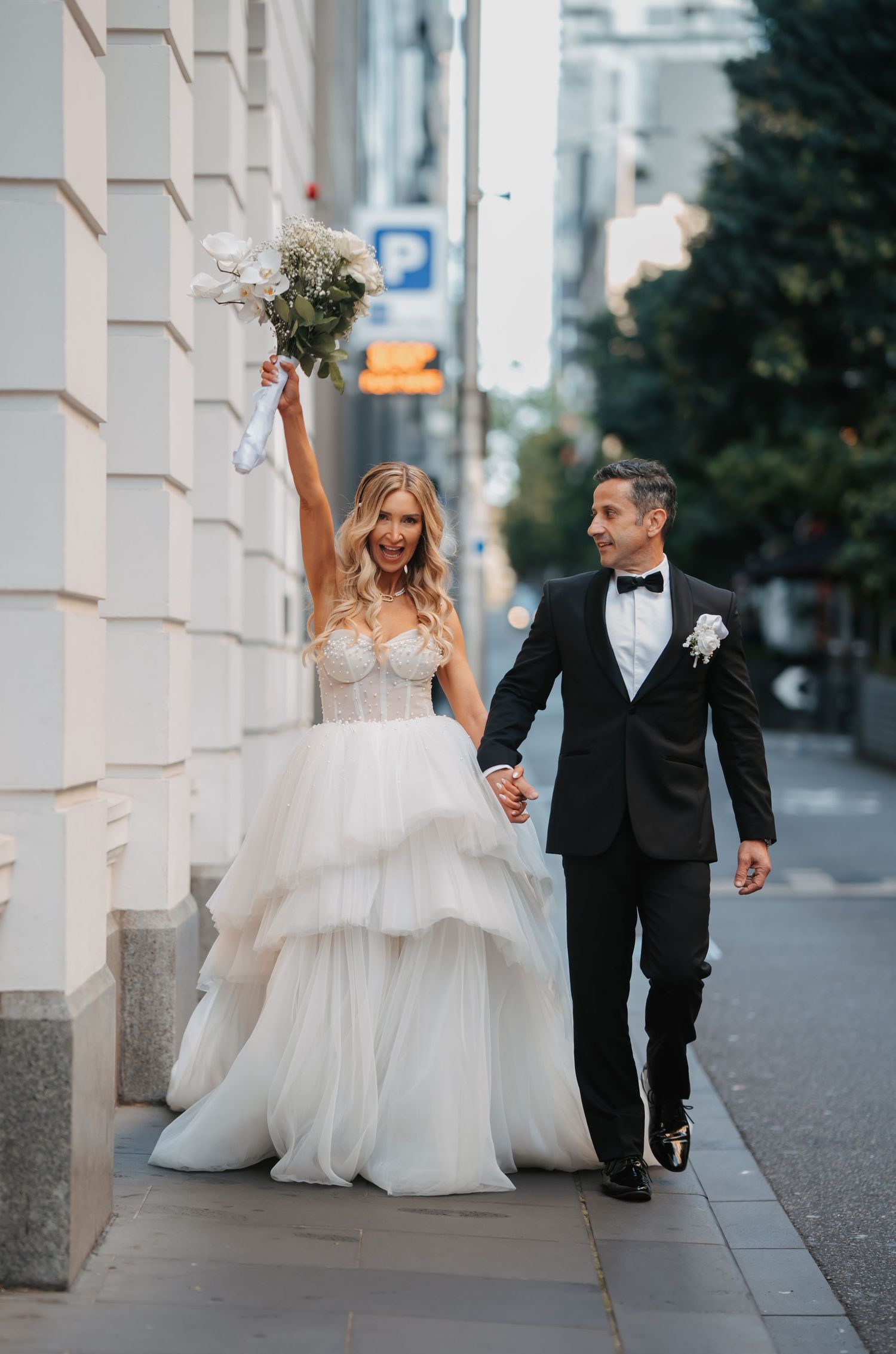 A couple in wedding attire walks and dances together on an urban sidewalk with white columns in the background.
