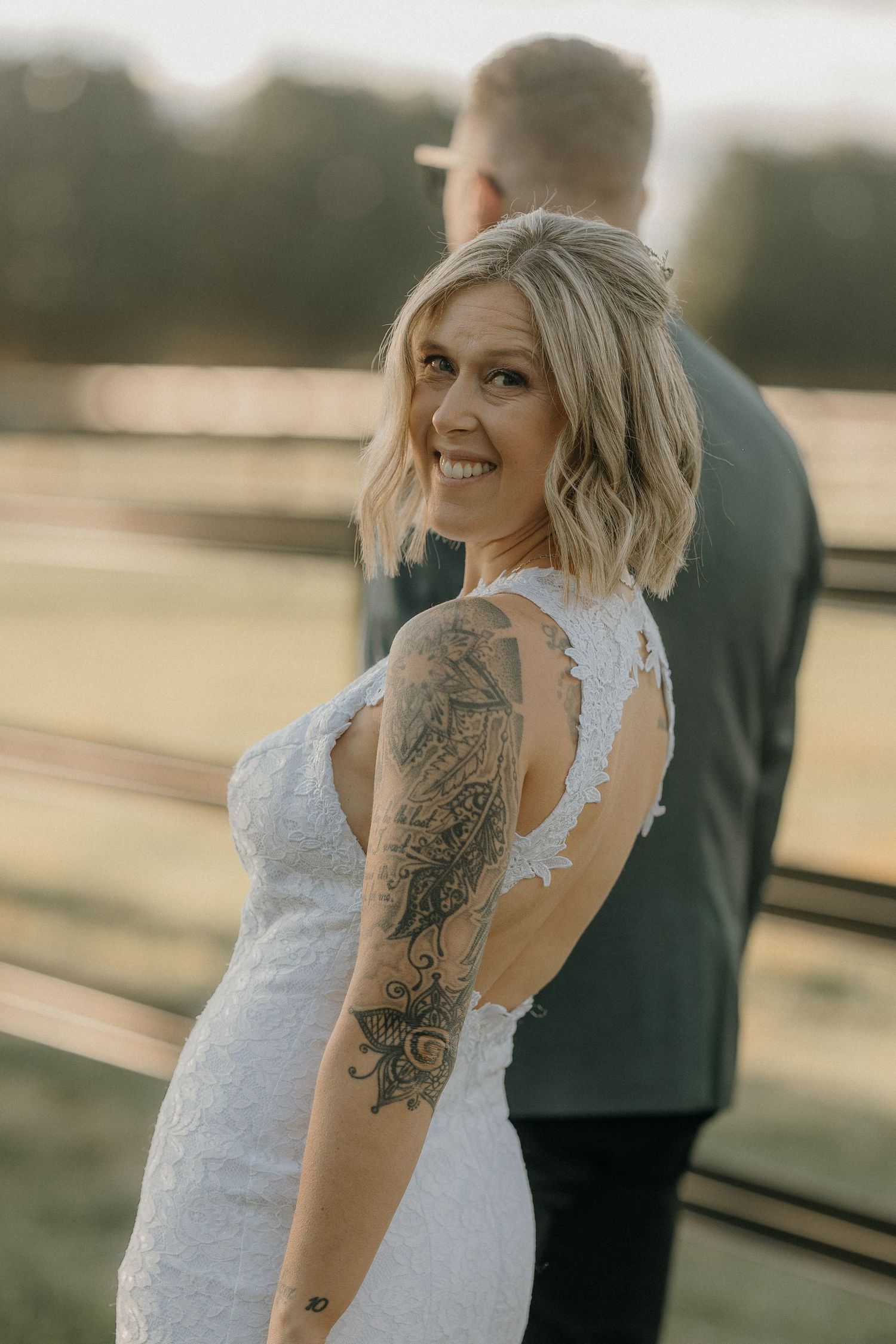 A bride in a white lace dress with visible tattoos smiles during an outdoor wedding photoshoot at sunset.