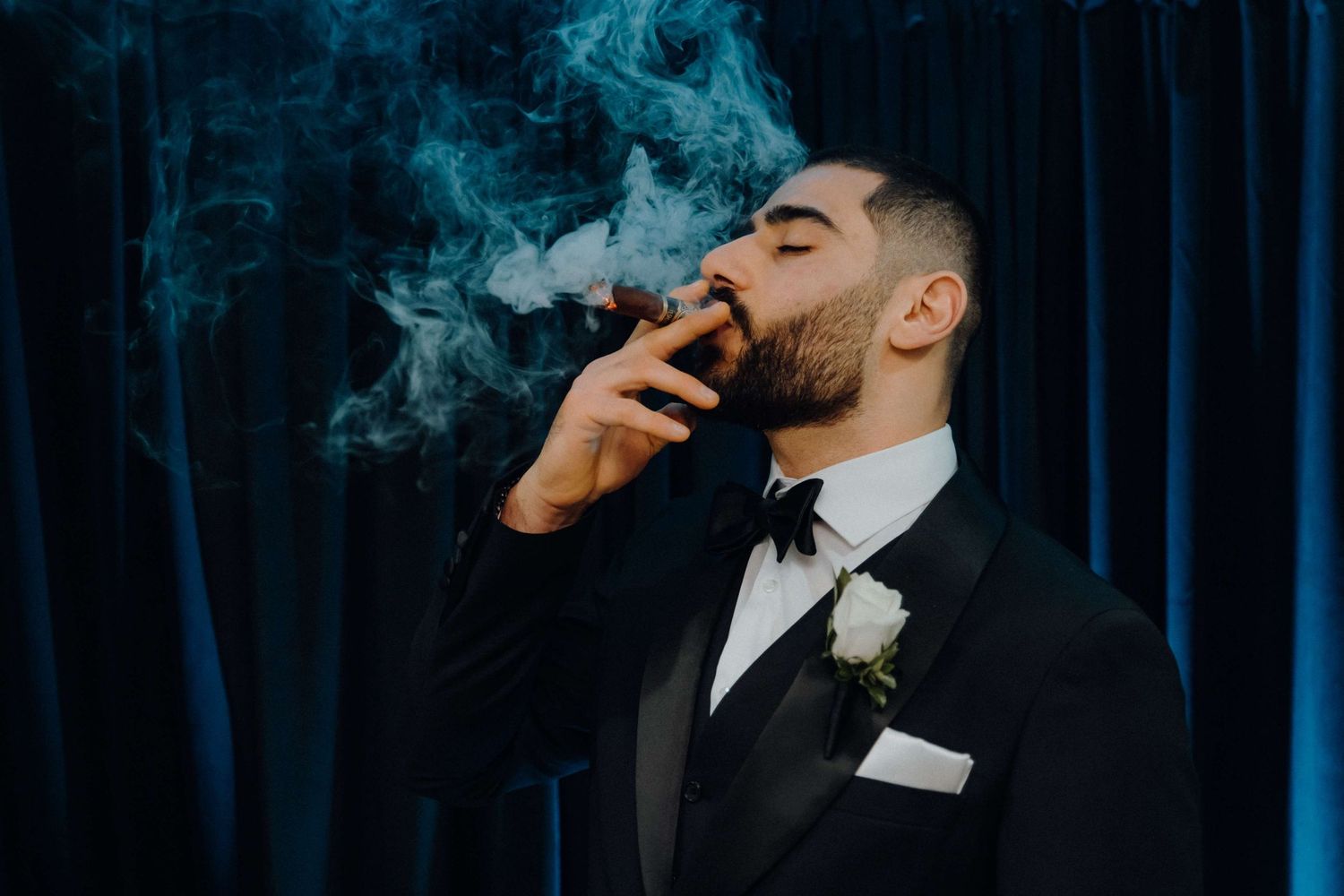 A groom in black tuxedo and white boutonniere exhales smoke against a dark blue curtain backdrop.