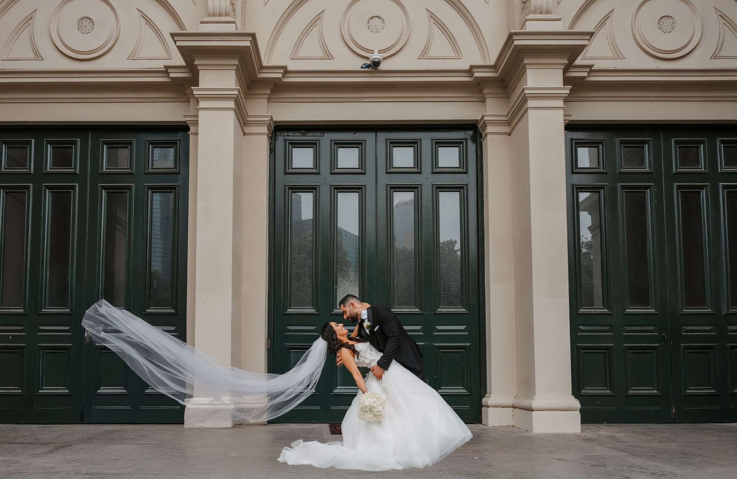 Elegant wedding photo showing a romantic embrace against majestic dark green doors while a veil flows dramatically in the breeze.