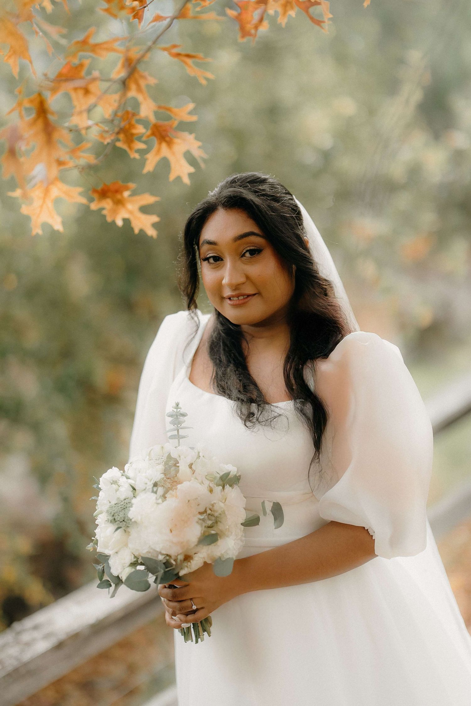 A bride in a white dress with sheer sleeves holds a delicate bouquet against an autumn backdrop with orange leaves.