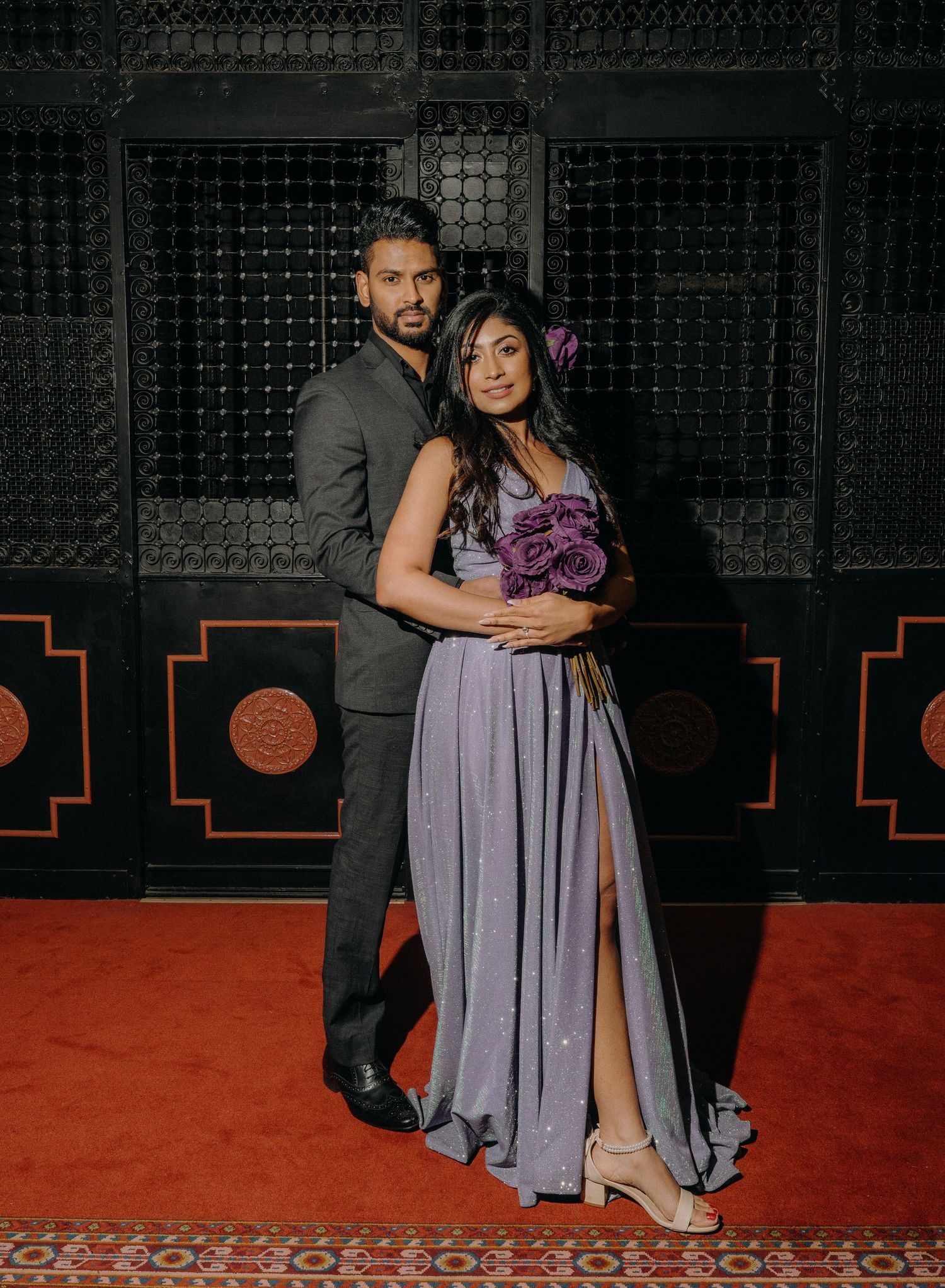 A couple poses against a dark background with dramatic lighting and a red carpet setting during what appears to be an event.