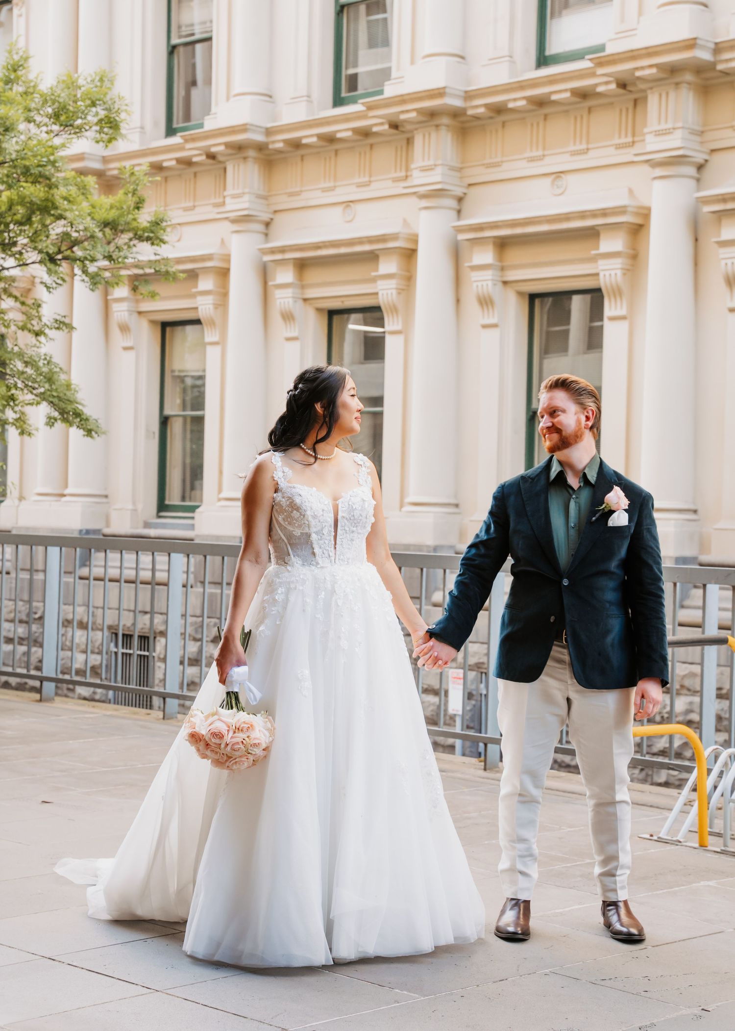 A bride in a white lace wedding gown and groom in a navy blazer holding hands outside a historic building.