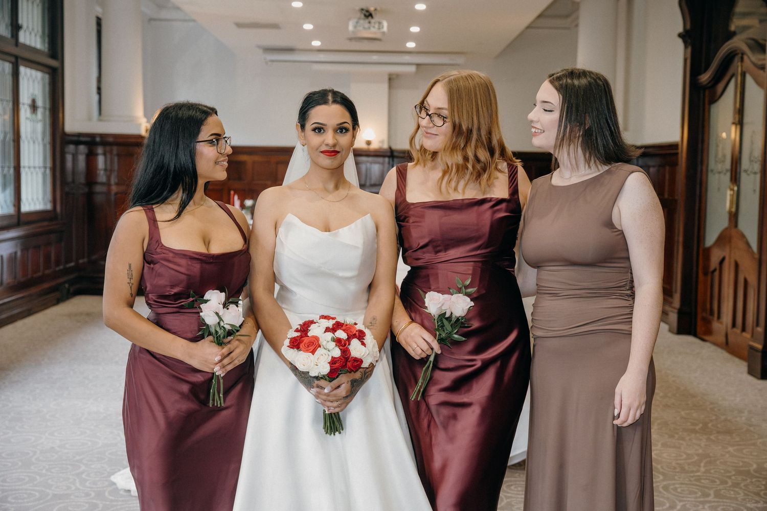 Bride in white gown with three bridesmaids in burgundy and taupe dresses holding red and white bouquets indoors.