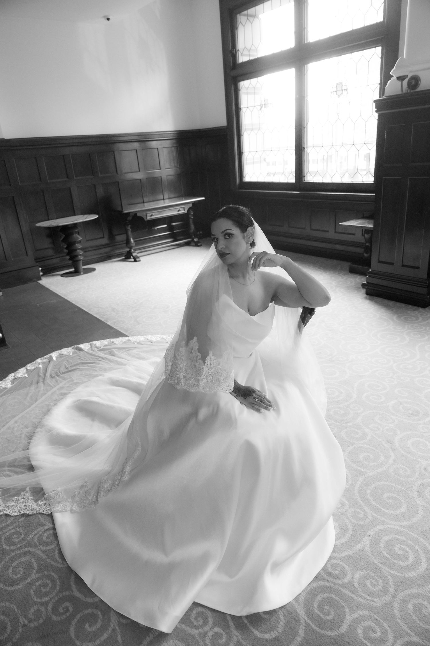 Bride in white gown and veil seated gracefully in elegant wood-paneled room, black and white wedding portrait.