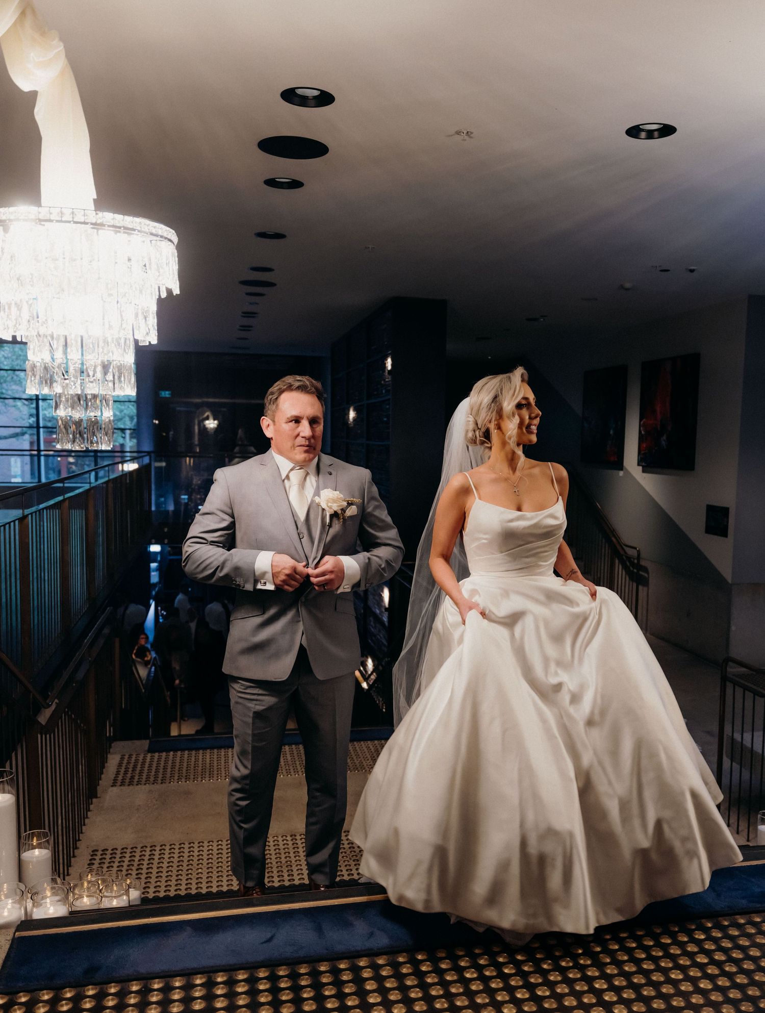 A couple walks down an elegant hallway with crystal chandeliers, with the bride wearing a stunning white ballgown.