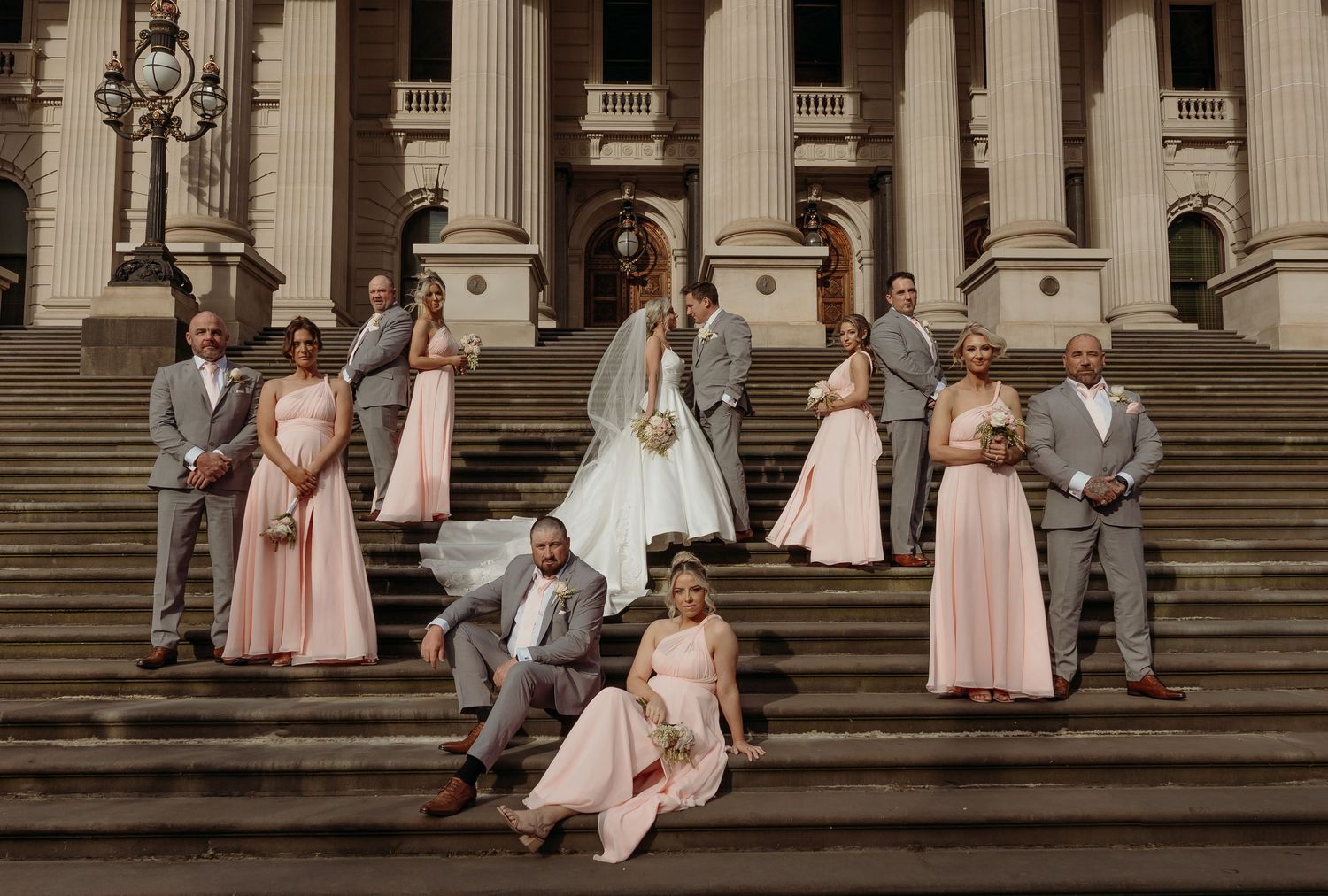 A wedding party in pink and gray attire poses elegantly on grand stone steps of a classical building with columns.