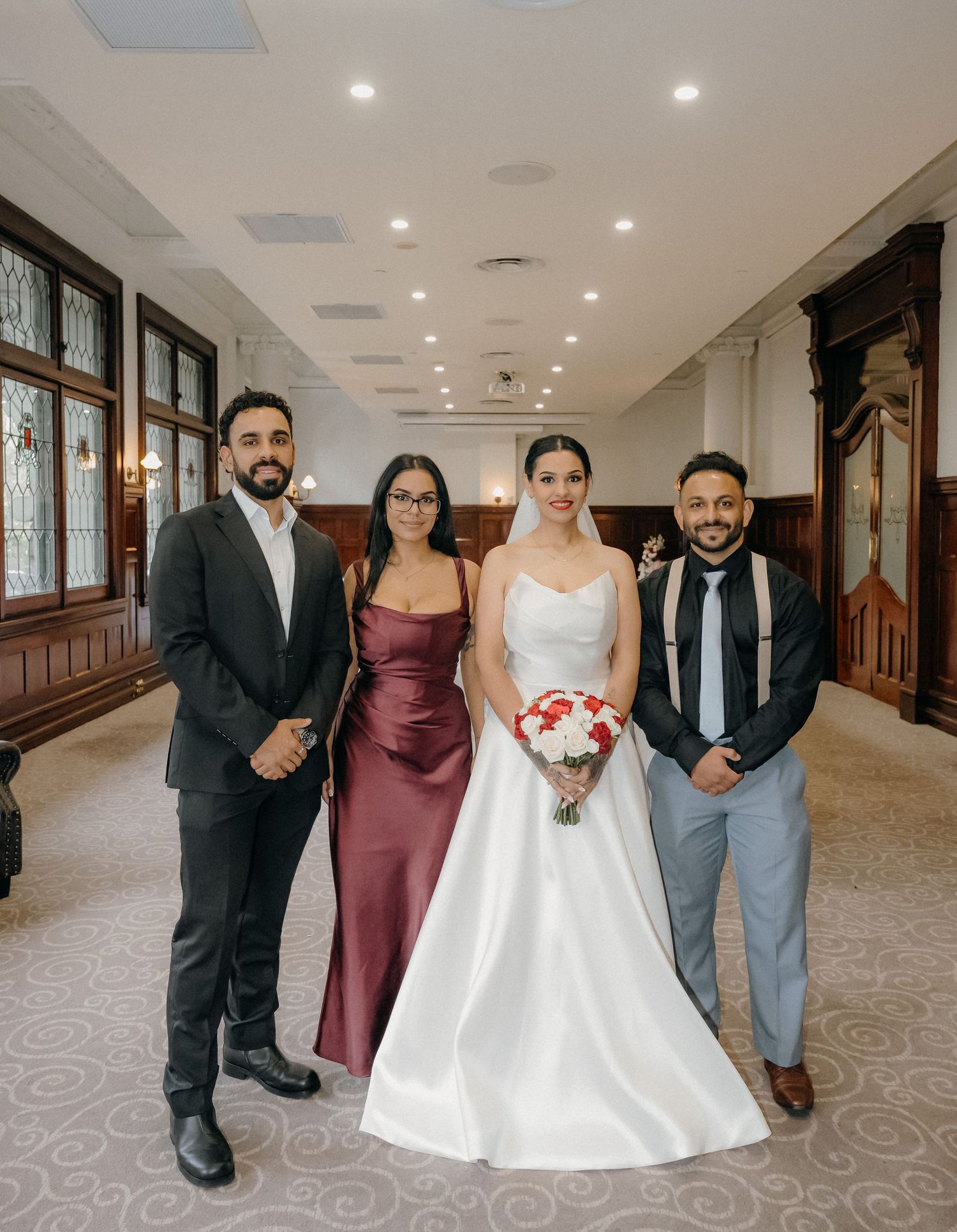 Bride in white gown holding bouquet poses with three guests in formal attire inside elegant wedding venue.