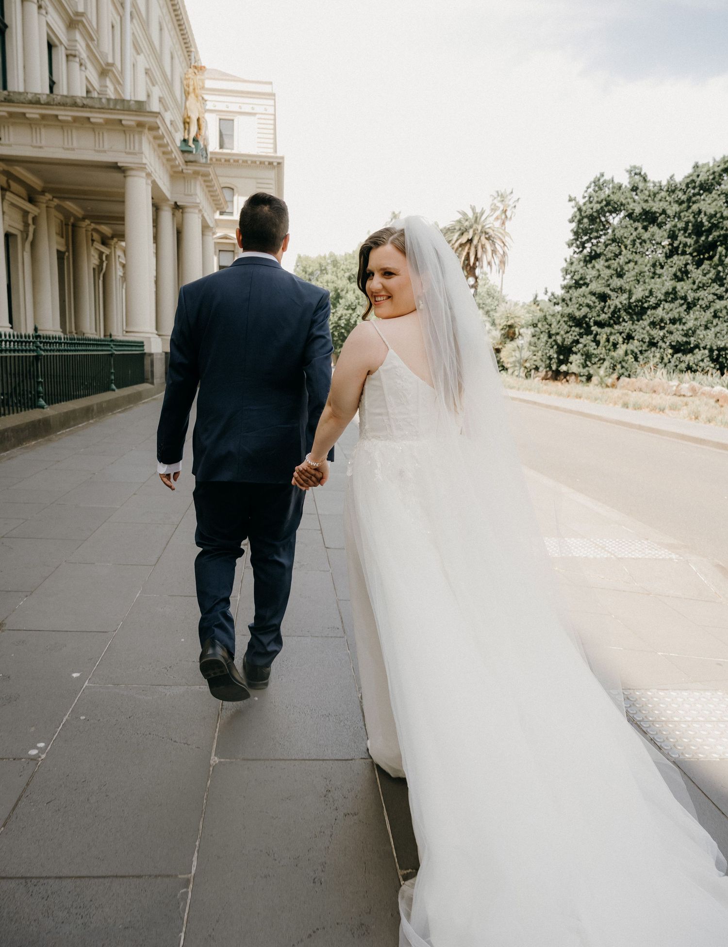 A couple in wedding attire walks down a grand outdoor colonnade in a series of progressive shots.
