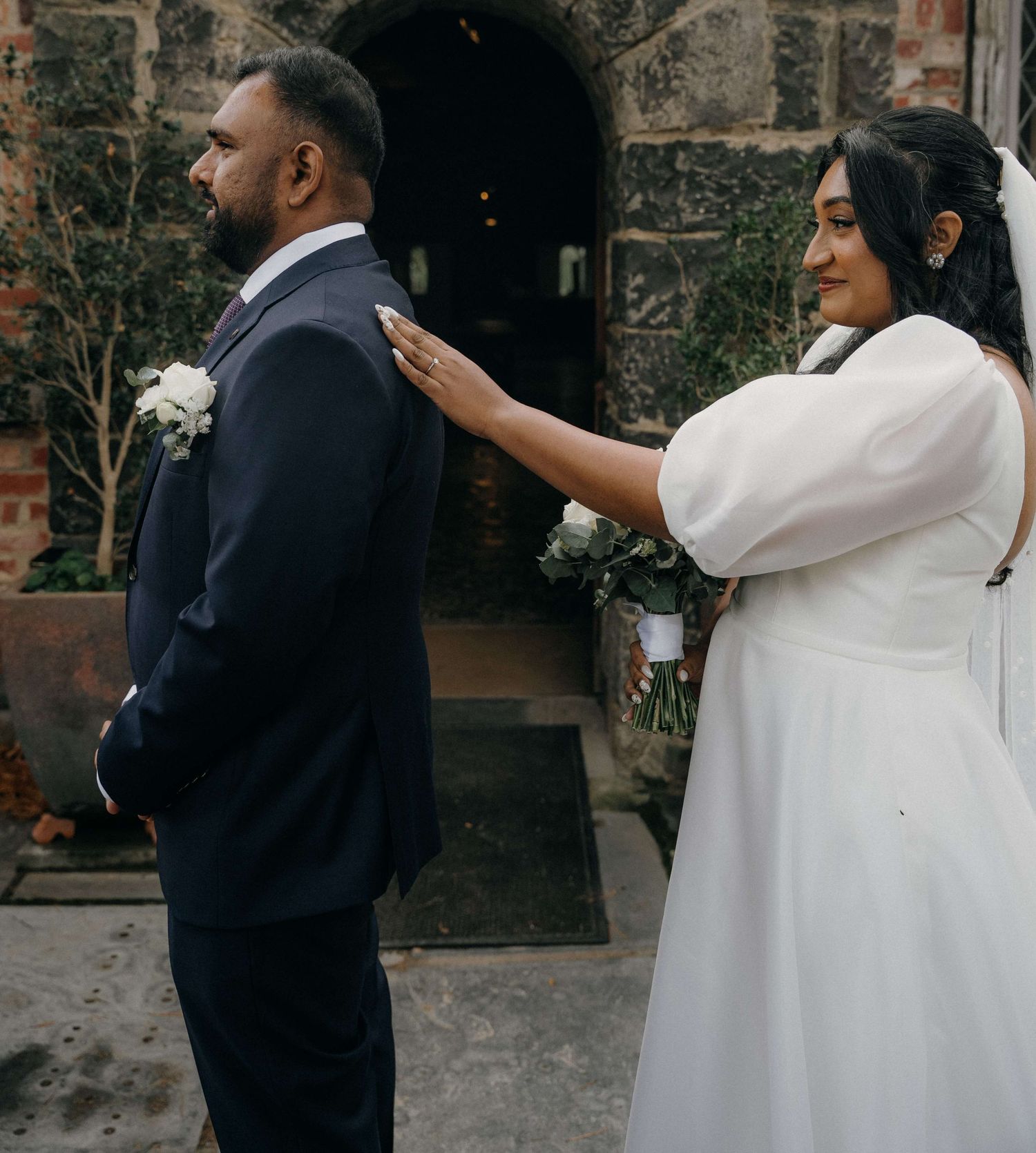 Elegant wedding couple shares a touching first look moment against a stone wall backdrop in classic formal attire.