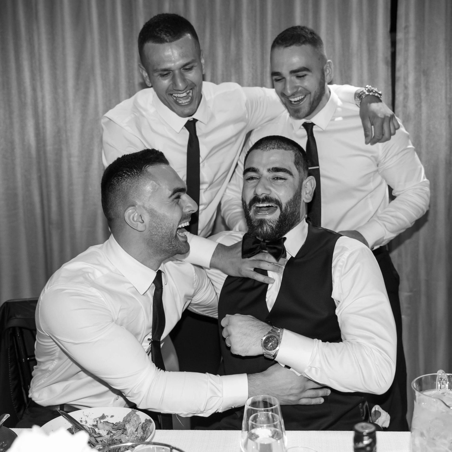 A black and white photo of four joyful men in formal attire laughing and embracing at a dinner table.