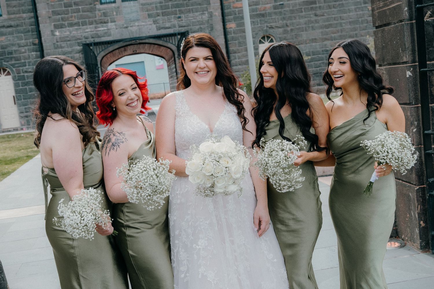 A group of bridesmaids wearing sage green dresses stand with a bride in white during a wedding celebration.
