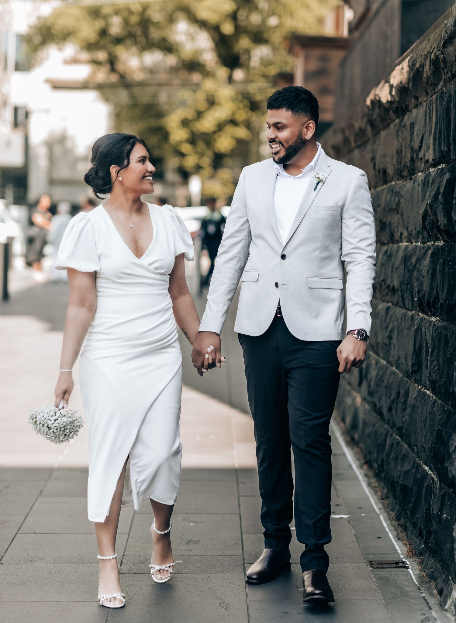 A couple in white wedding attire walks hand in hand along a stone wall on their special day.