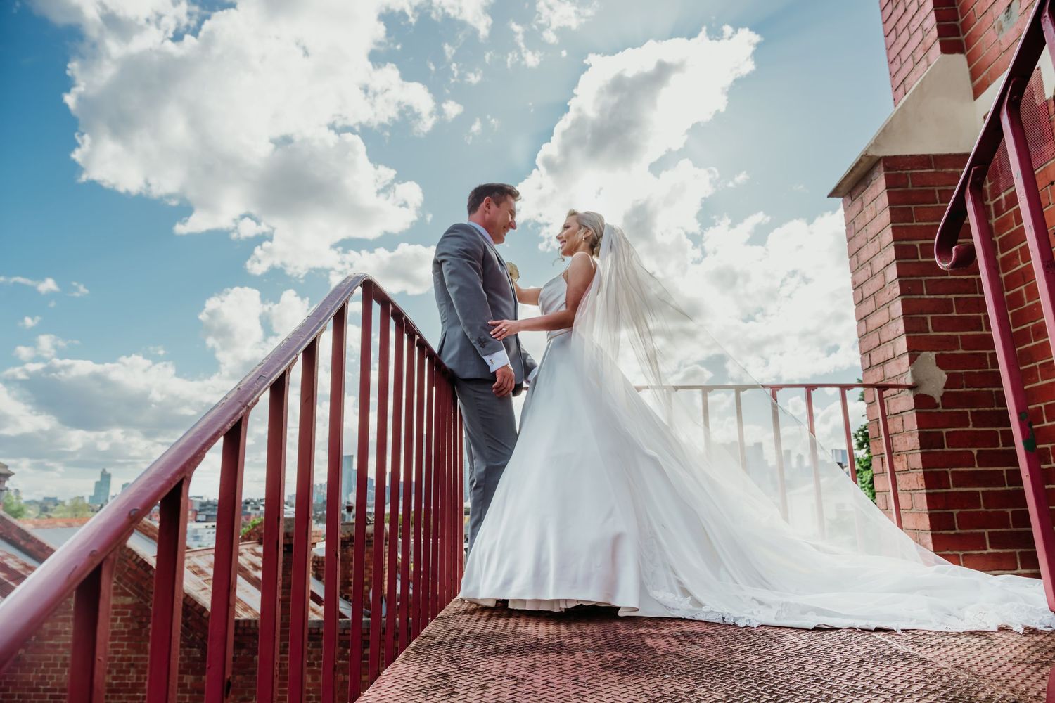 A couple in wedding attire stands together on red brick stairs with a metal railing against a blue cloudy sky.
