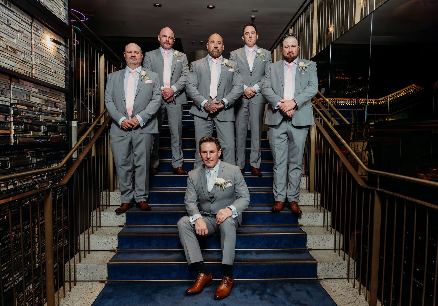Wedding party in light gray suits poses on elegant navy blue staircase with gold railings in sequence of five photos.