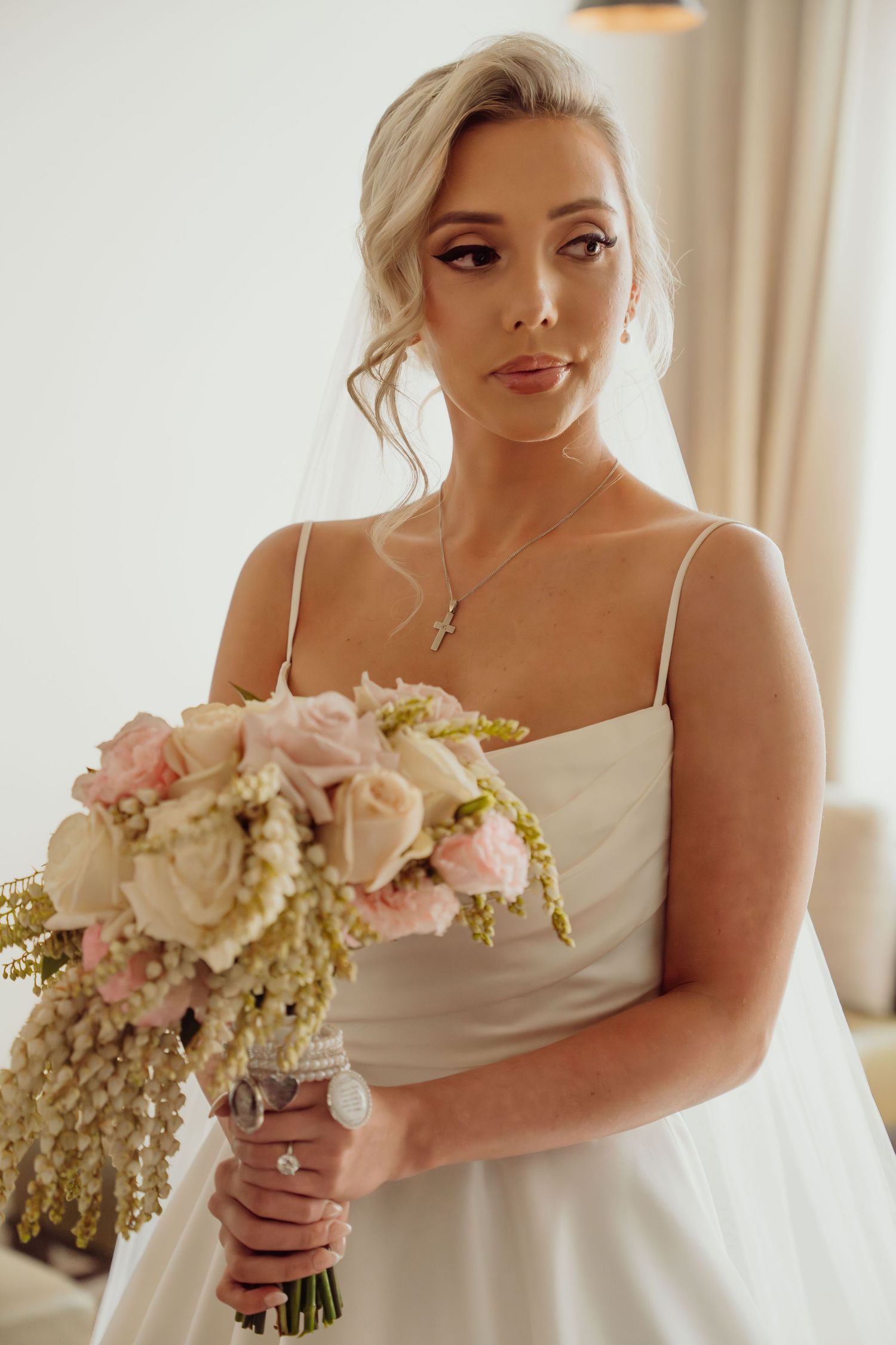 Bride in white wedding dress holds elegant bouquet of blush and ivory flowers in sunlit room.