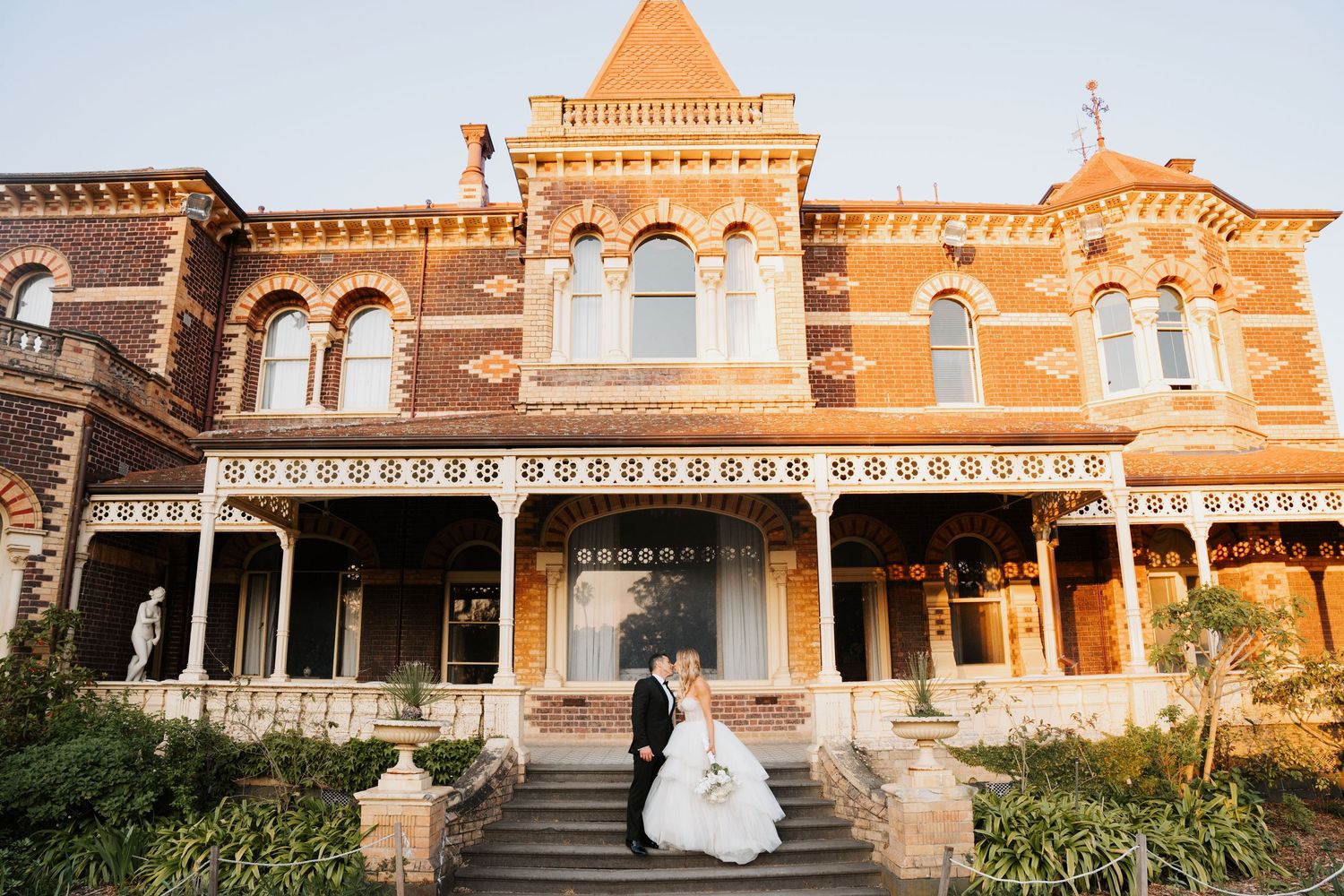 A wedding couple stands in front of a grand Victorian-style mansion with ornate architecture at sunset.
