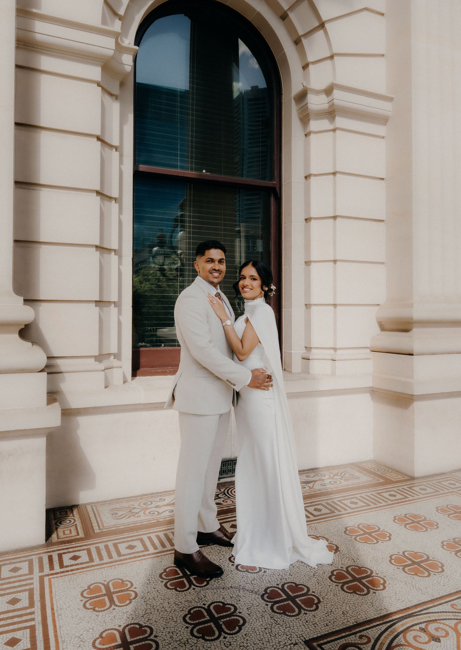 A couple in white attire poses elegantly against ornate columns with decorative floor tiles at a historic building.