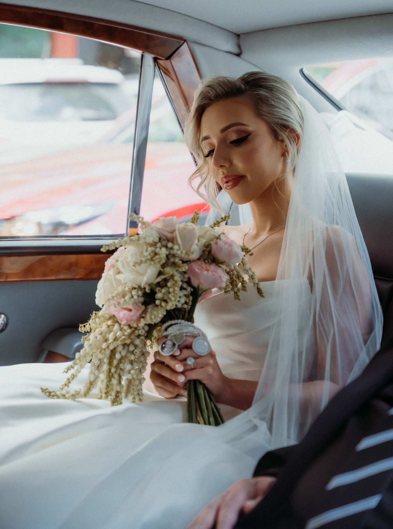 Bride in wedding dress holding a bouquet of pink and white flowers while seated in a vintage car.
