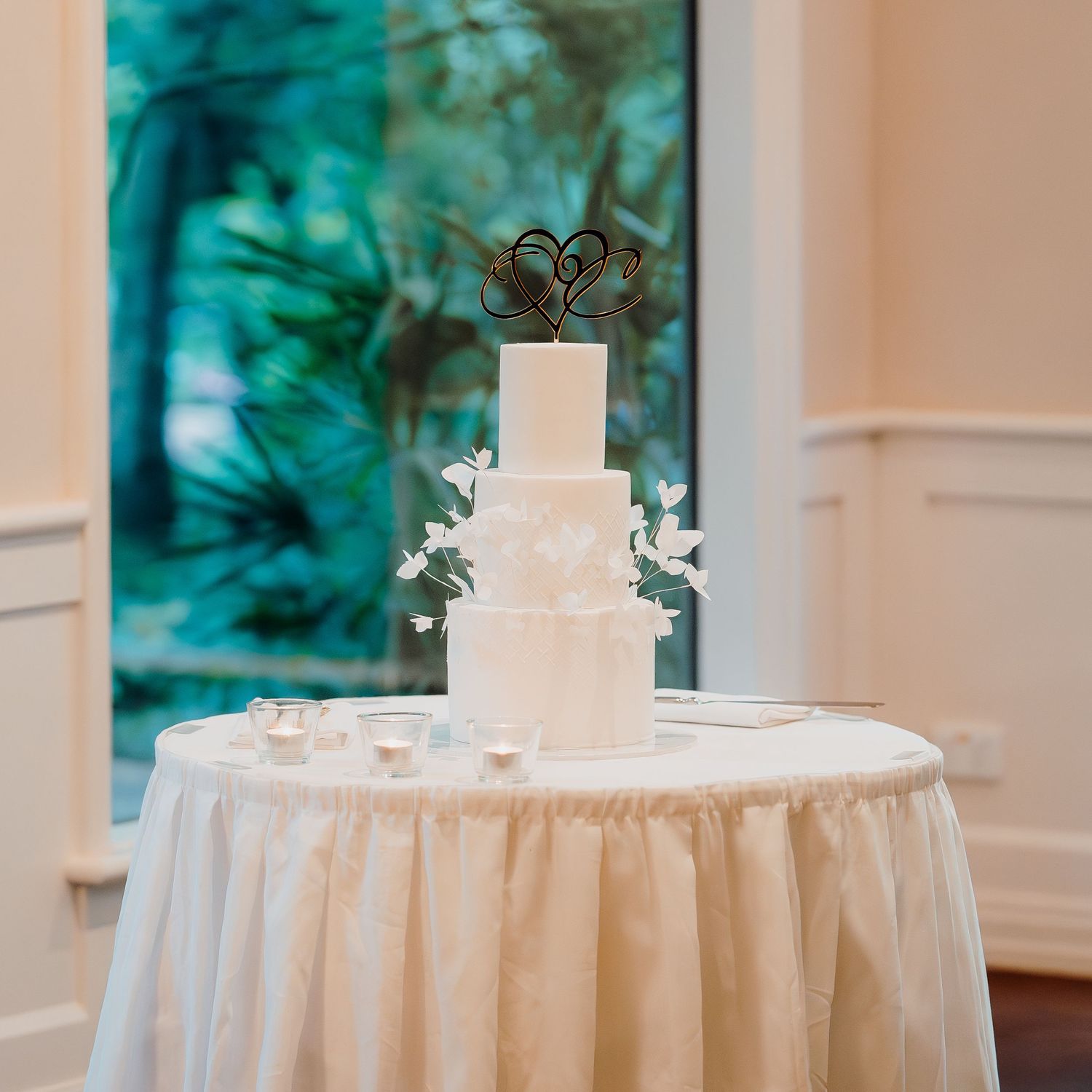Elegant white wedding cake displayed on round table with white tablecloth and floral decorations in a bright venue.