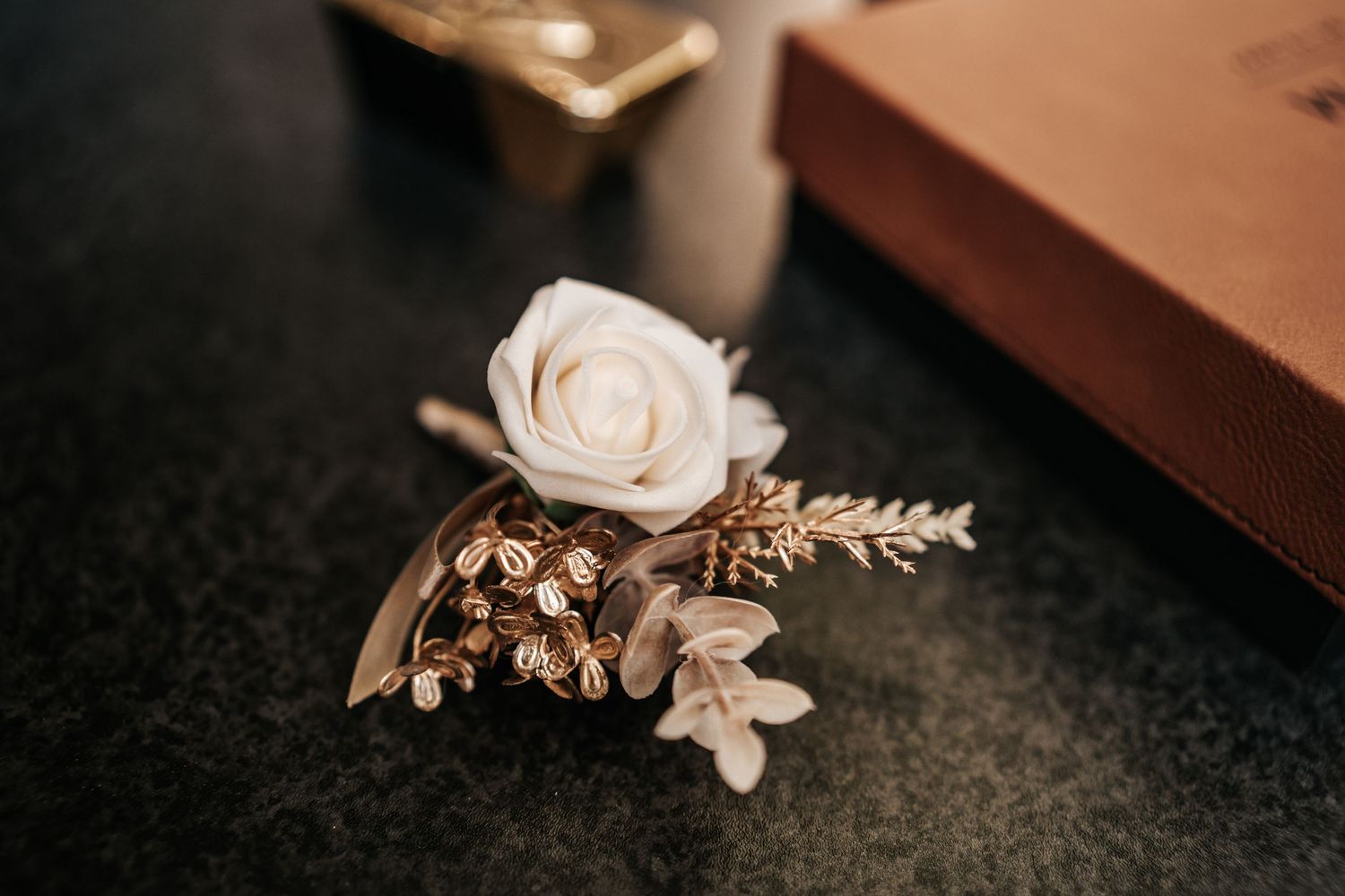 A delicate cream rose boutonniere adorned with small gold leaves and floral accents rests on a dark surface.