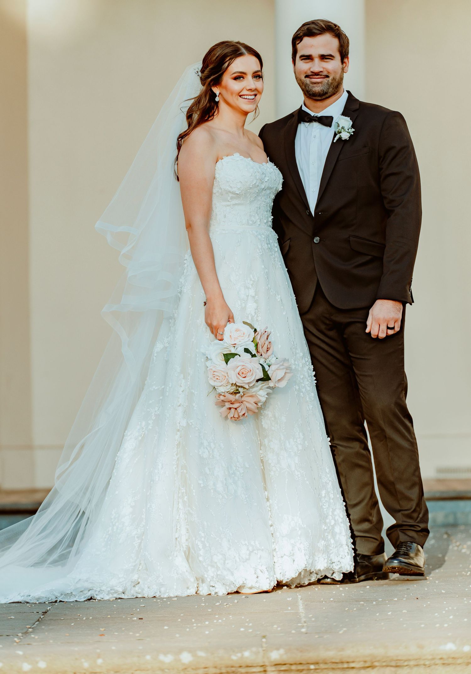 A newlywed couple poses together in formal wedding attire with a bouquet in front of a light-colored wall.
