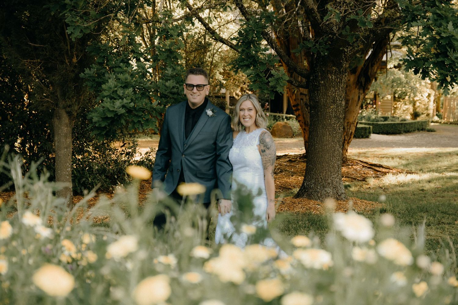 A couple stands together on a path in a park surrounded by trees and delicate white wildflowers on a sunny day.