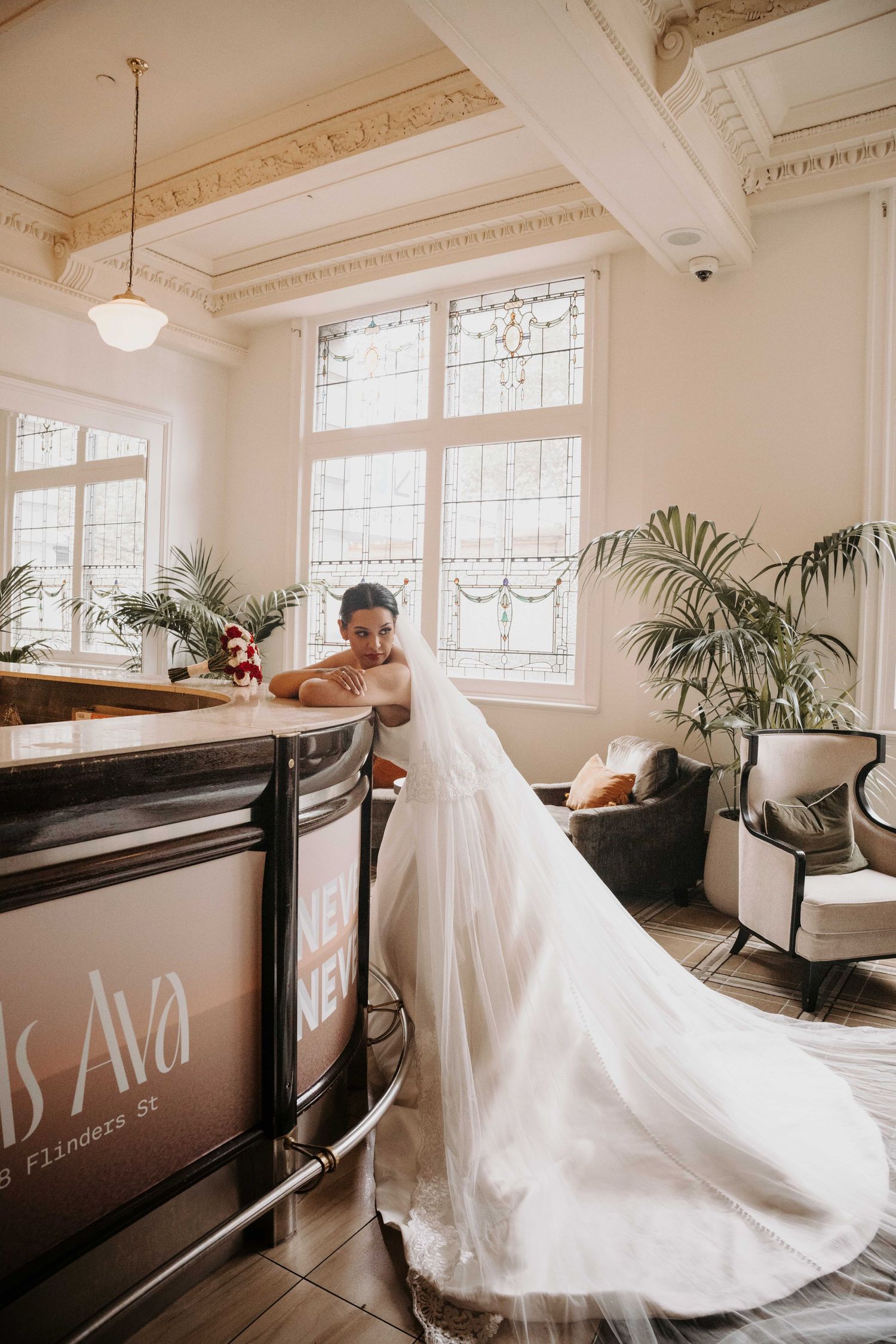 Bride in a flowing white gown and veil leans against a bar counter in an elegant venue with stained glass windows.