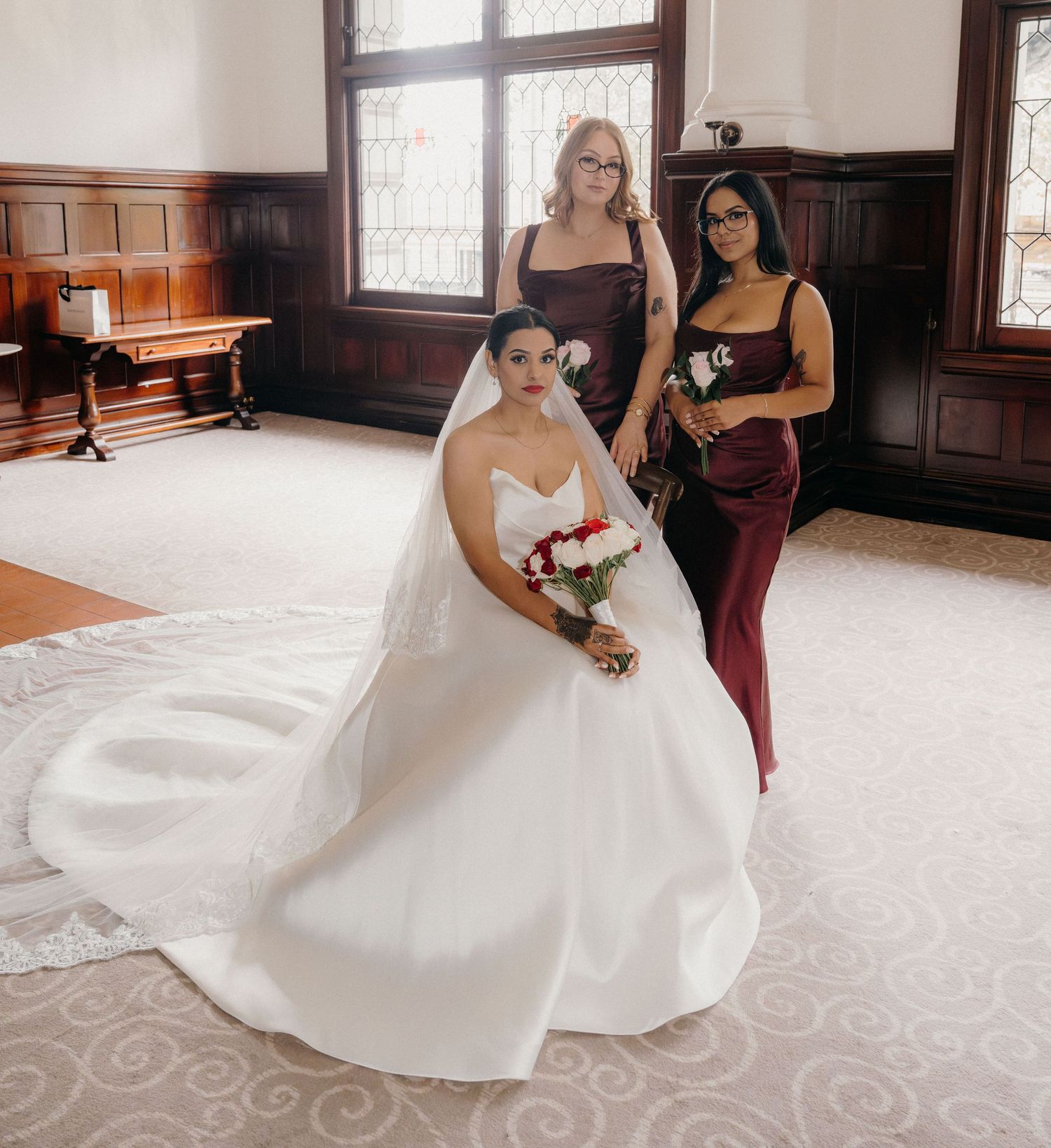 Bride in white ball gown with veil holds red bouquet, flanked by two bridesmaids in burgundy dresses.