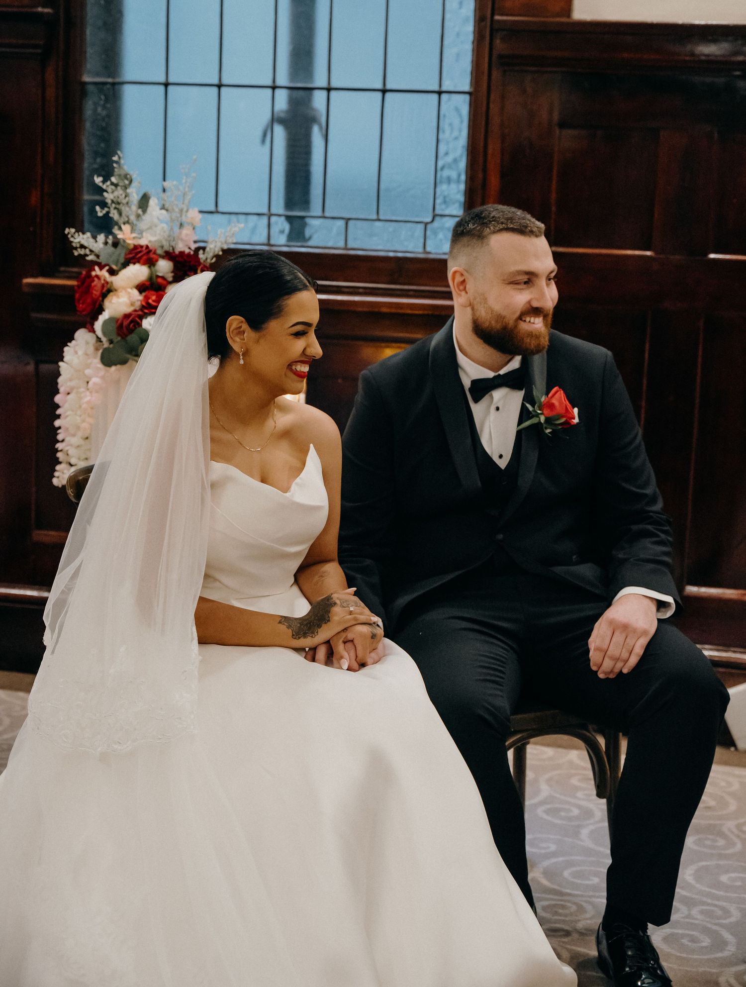Bride in white strapless gown and groom in black tuxedo sitting together smiling during wedding ceremony.
