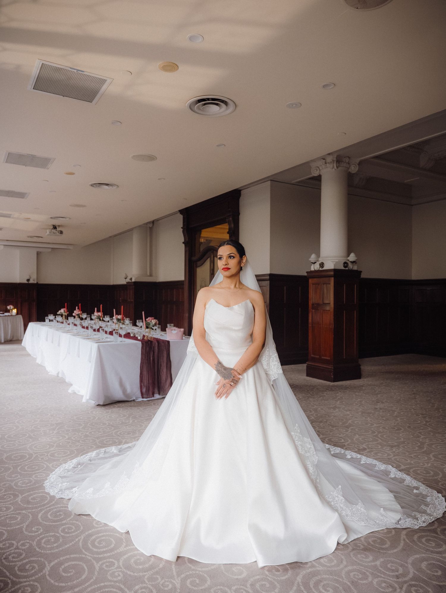 Bride in a strapless white ball gown with a long train poses in an elegant wedding reception hall with draped tables.