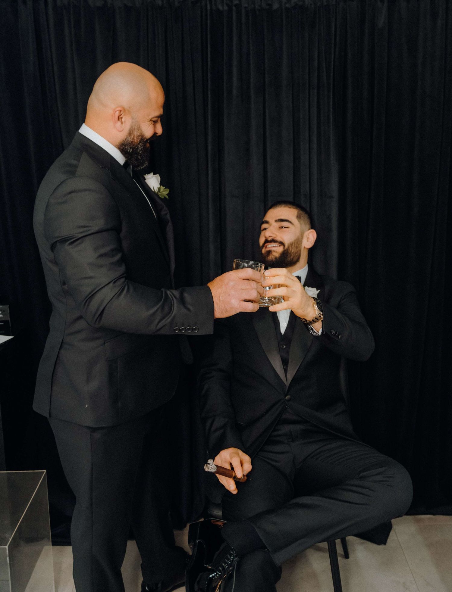 Two men in black suits share a toast and laugh together against a dark backdrop during a formal occasion.