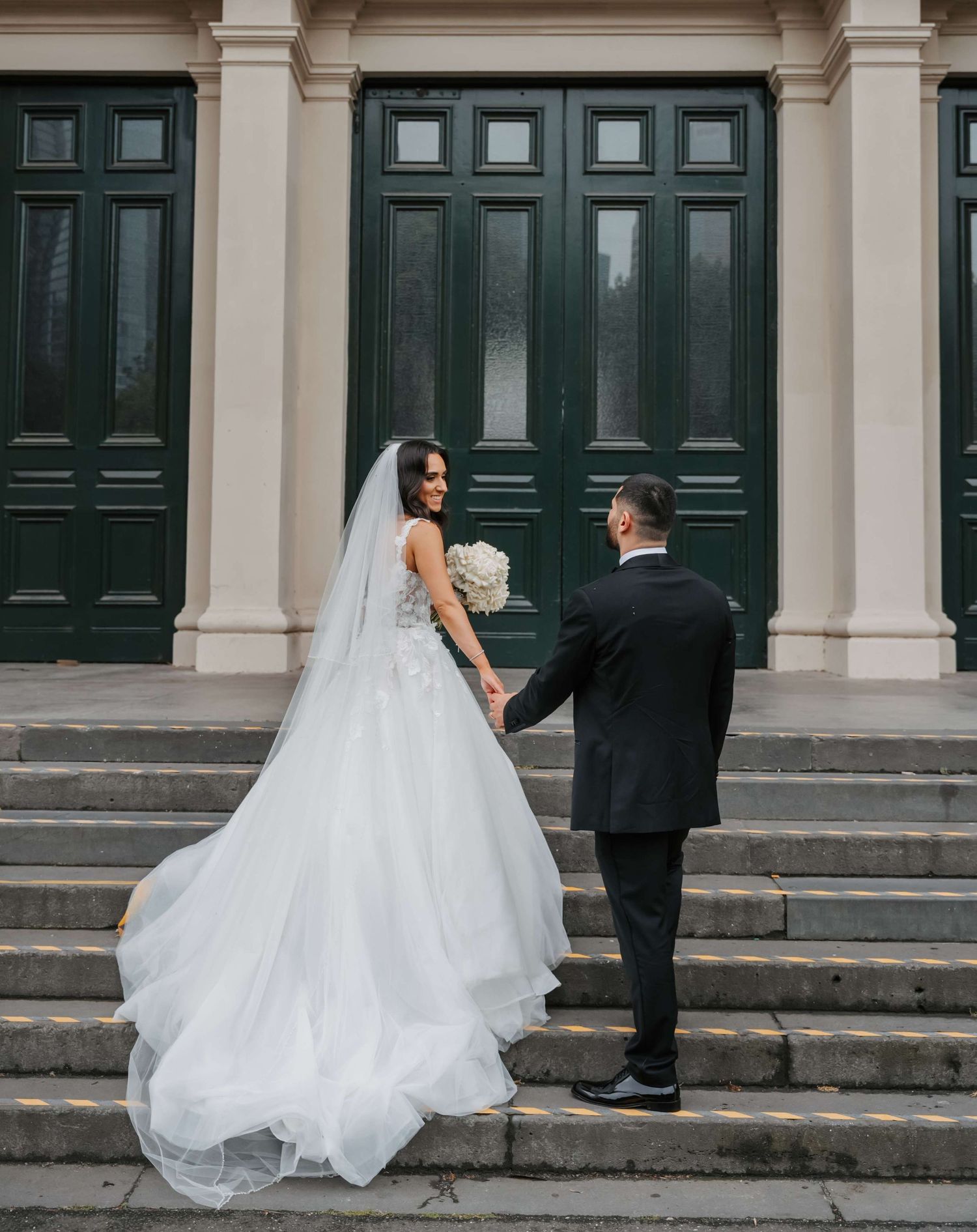 Newlyweds walk up grand stone steps with tall dark green doors and cream columns of a historic building.